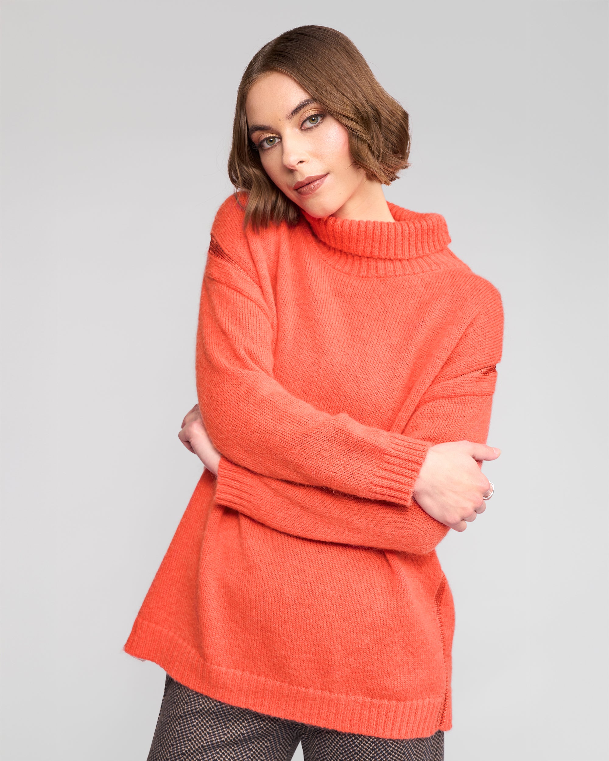 A woman with short brown hair models the Vassalli Mellow Sweater - Lava, an oversized orange turtleneck, paired with patterned pants. She poses with arms crossed and head tilted against a plain light gray background.