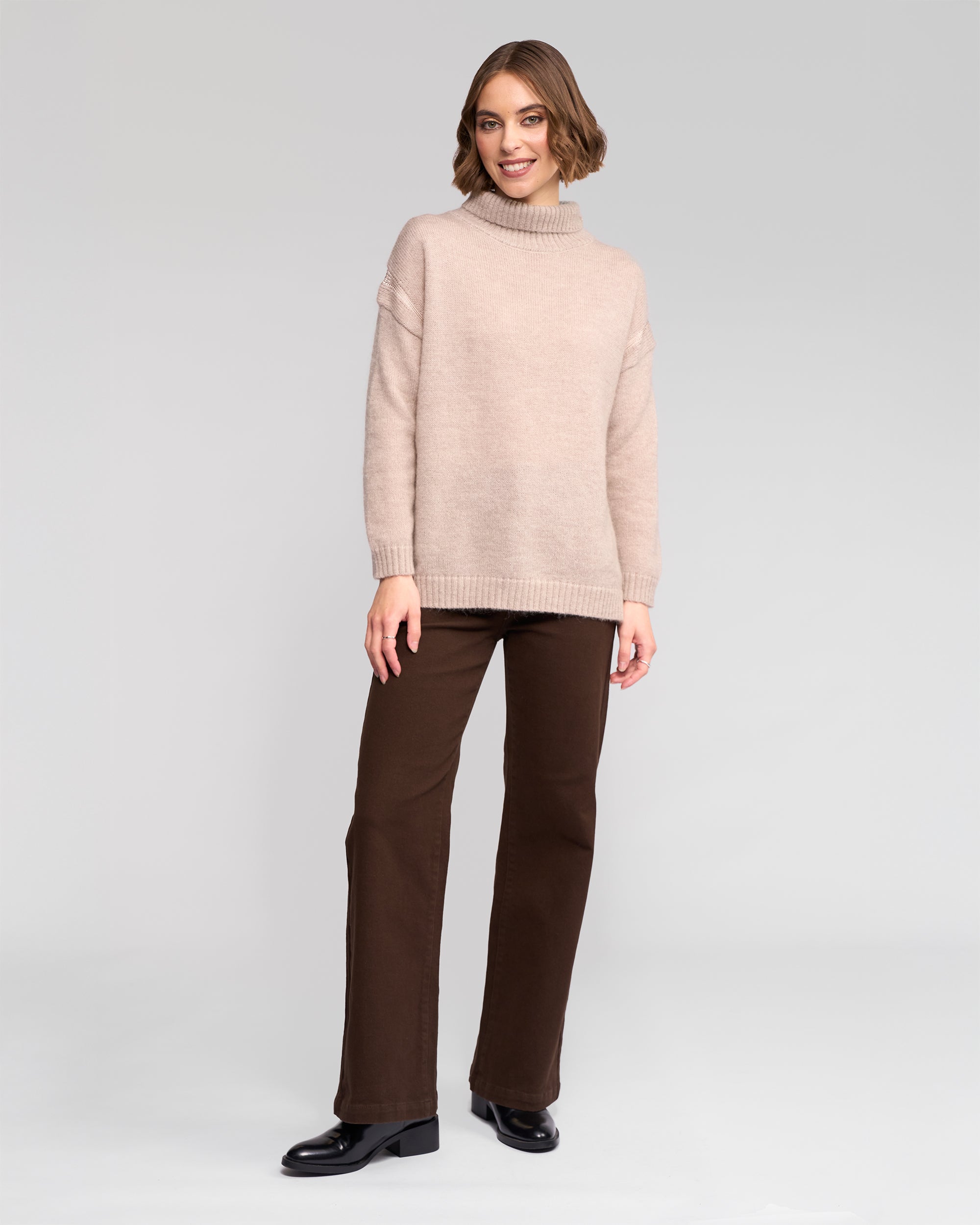A woman stands smiling in the Vassalli Mellow Sweater - Oat, paired with brown pants and black shoes, against a plain light gray background.