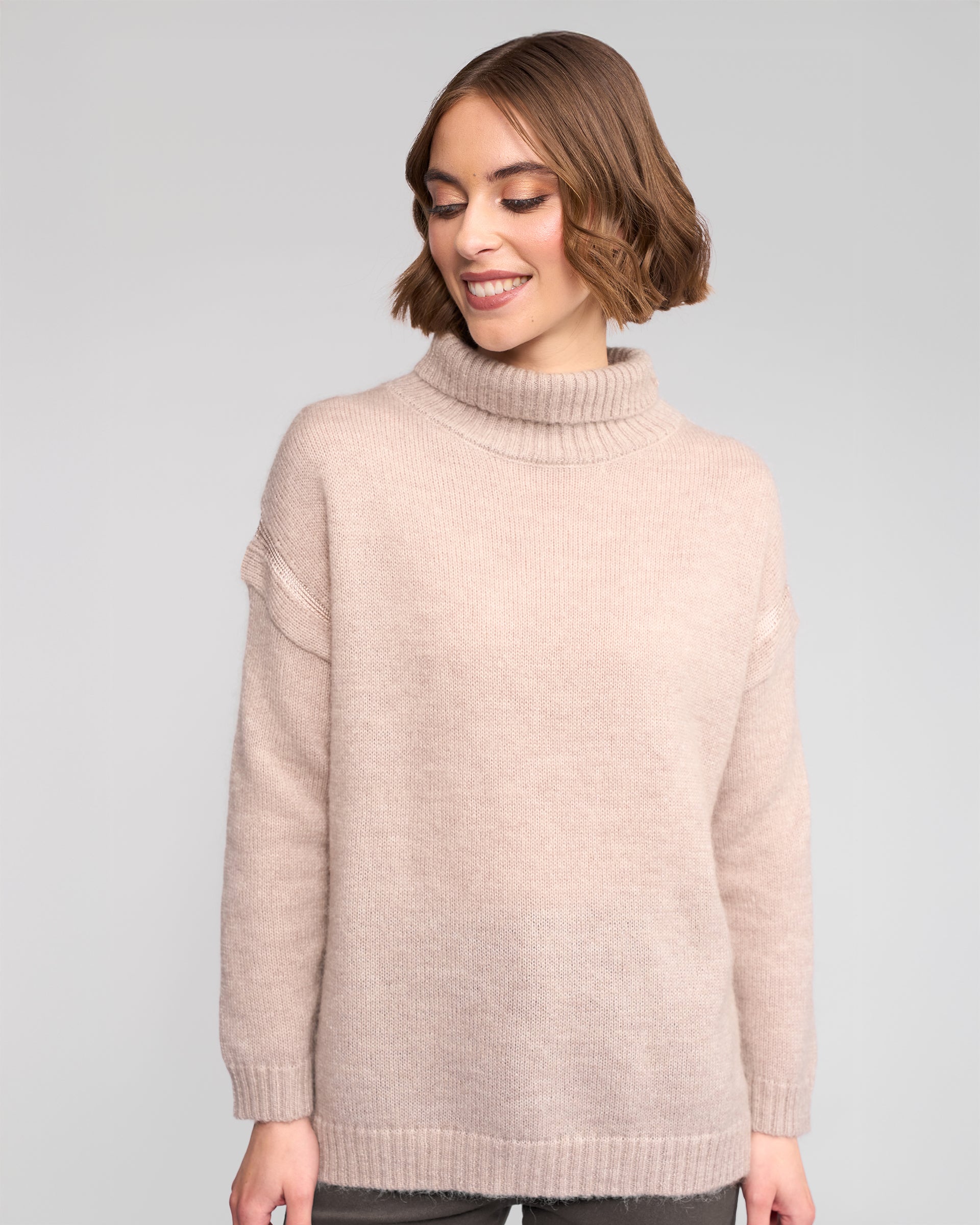 A woman with short brown hair wears the Vassalli Mellow Sweater in Oat, standing against a plain gray background, looking down and smiling.
