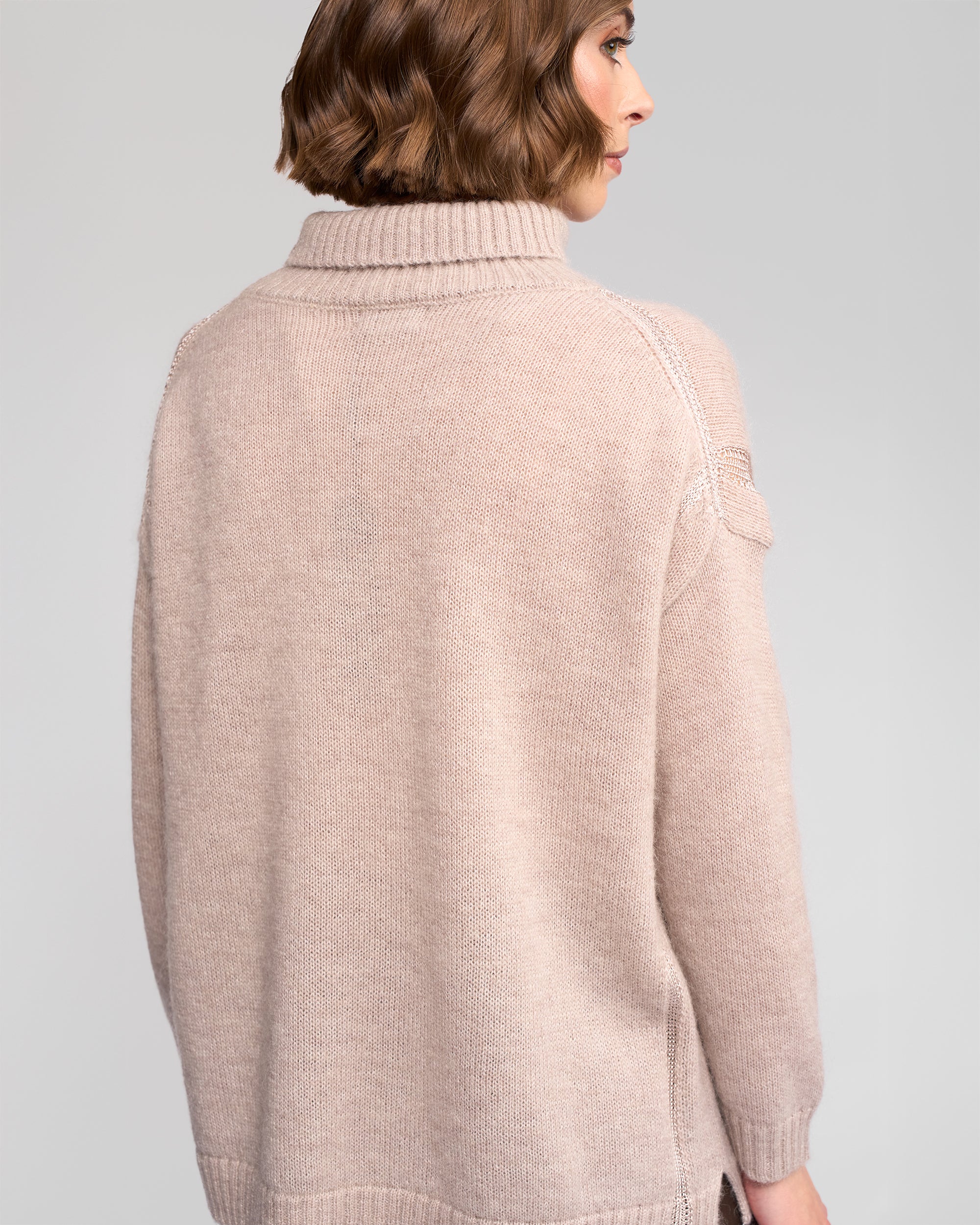 A woman with short wavy brown hair is seen from behind, wearing the Vassalli Mellow Sweater - Oat, an oversized beige mohair wool piece with long sleeves and a ribbed neck, set against a plain light background.
