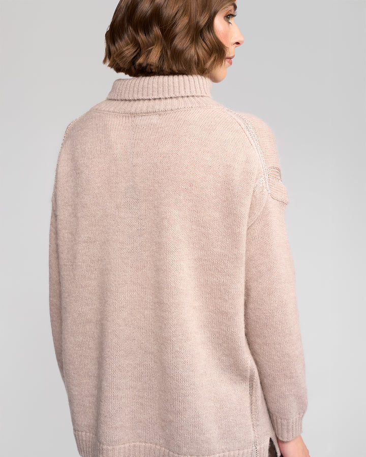A woman with short wavy brown hair is seen from behind, wearing the Vassalli Mellow Sweater - Oat, an oversized beige mohair wool piece with long sleeves and a ribbed neck, set against a plain light background.