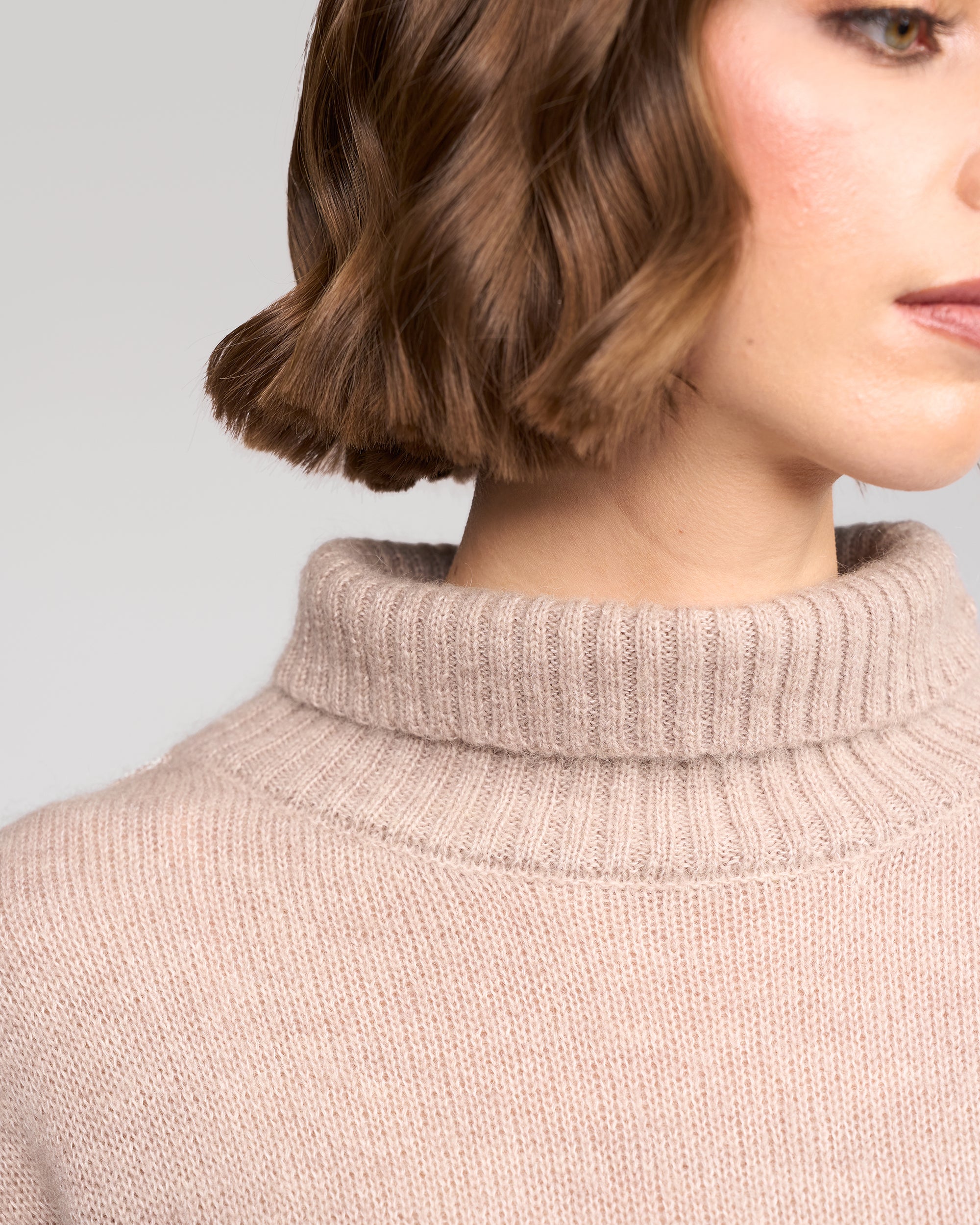 A close-up of a woman wearing the Vassalli Mellow Sweater in Oat, showing only the lower half of her face, neck, and shoulder. Her short brown hair is softly waved.