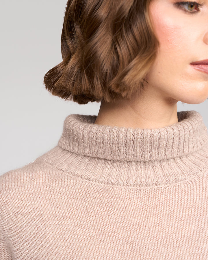 A close-up of a woman wearing the Vassalli Mellow Sweater in Oat, showing only the lower half of her face, neck, and shoulder. Her short brown hair is softly waved.
