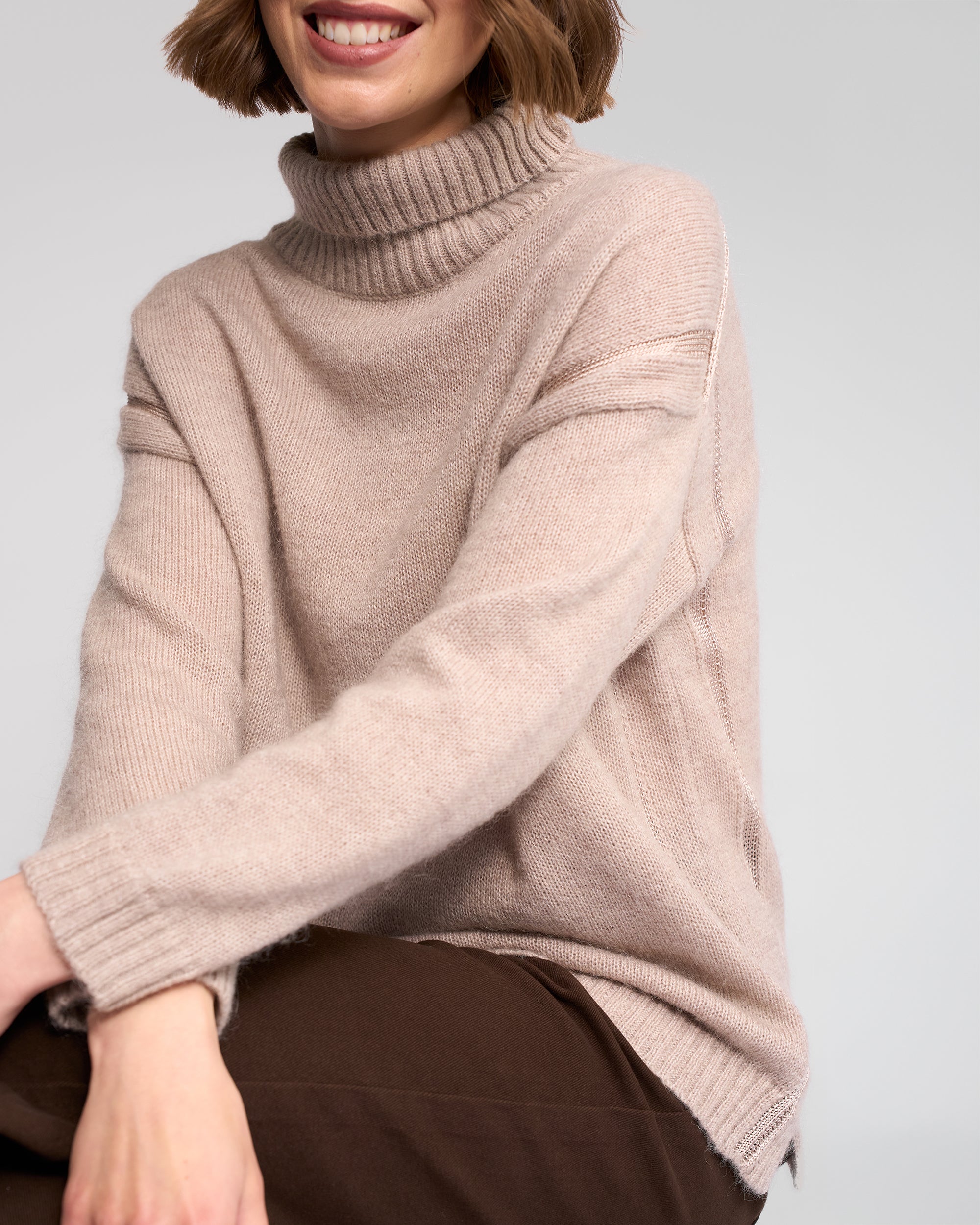 A person with short brown hair is smiling while sitting against a light gray background, wearing the Vassalli Mellow Sweater in Oat. Their face is partially out of frame, and they pair the look with dark pants.