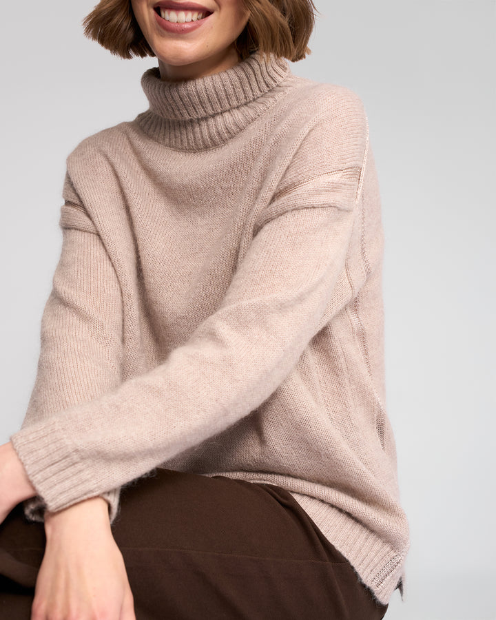 A person with short brown hair is smiling while sitting against a light gray background, wearing the Vassalli Mellow Sweater in Oat. Their face is partially out of frame, and they pair the look with dark pants.