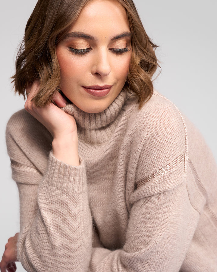 A woman with wavy brown hair wears the Vassalli Mellow Sweater in Oat, gazing down gently with her chin resting on her hand against a neutral background.