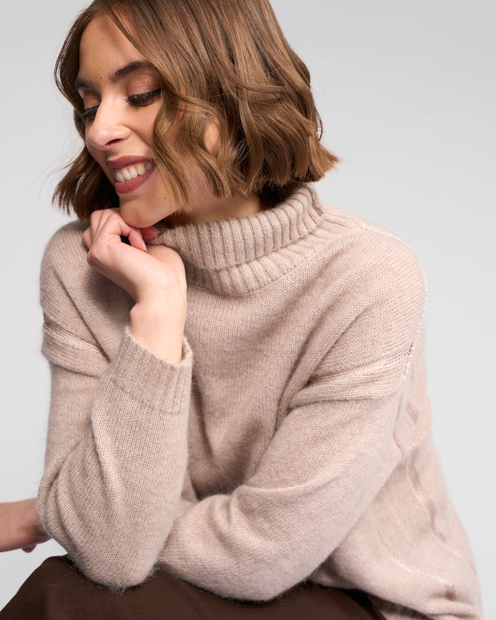 A woman with short, wavy brown hair smiles while looking down. She wears the Vassalli Mellow Sweater - Oat, an oversized beige knit with a ribbed neck, and rests one hand near her chin against a plain light background.