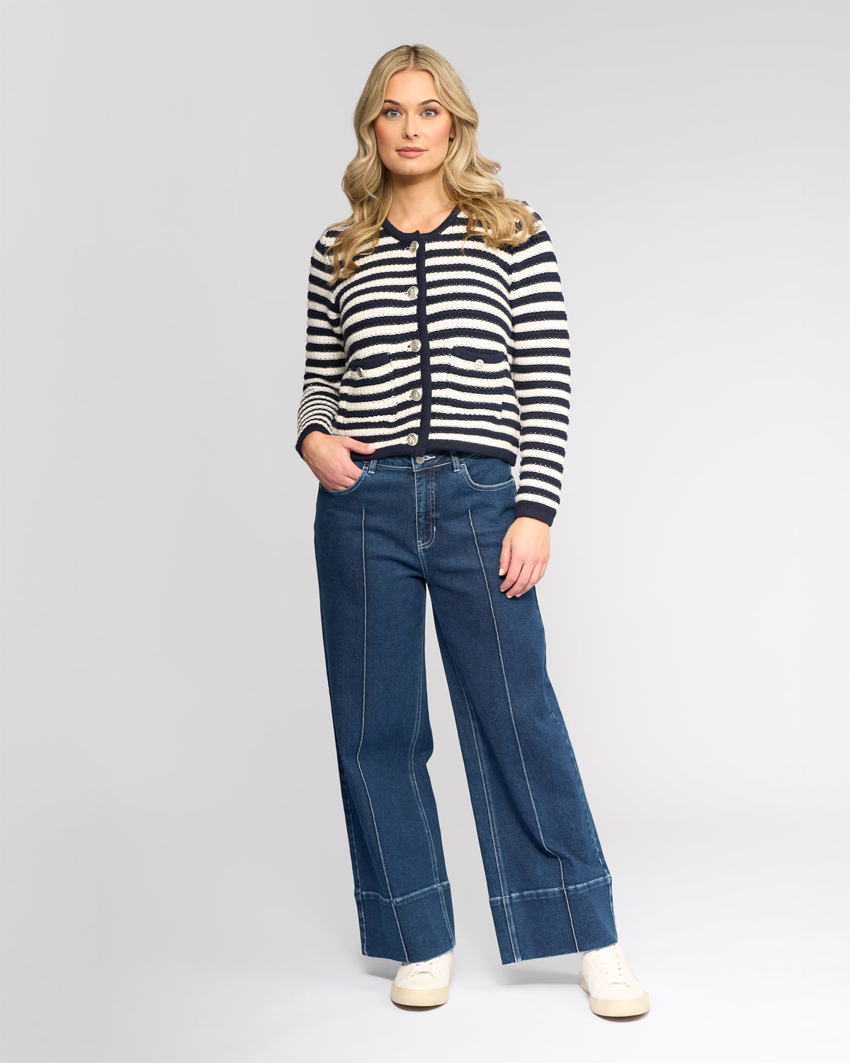 A woman with long blonde hair wears the Vassalli Avenue Cardi in Ink/Cream, styled with wide-leg blue jeans and white sneakers, standing against a plain light gray background.