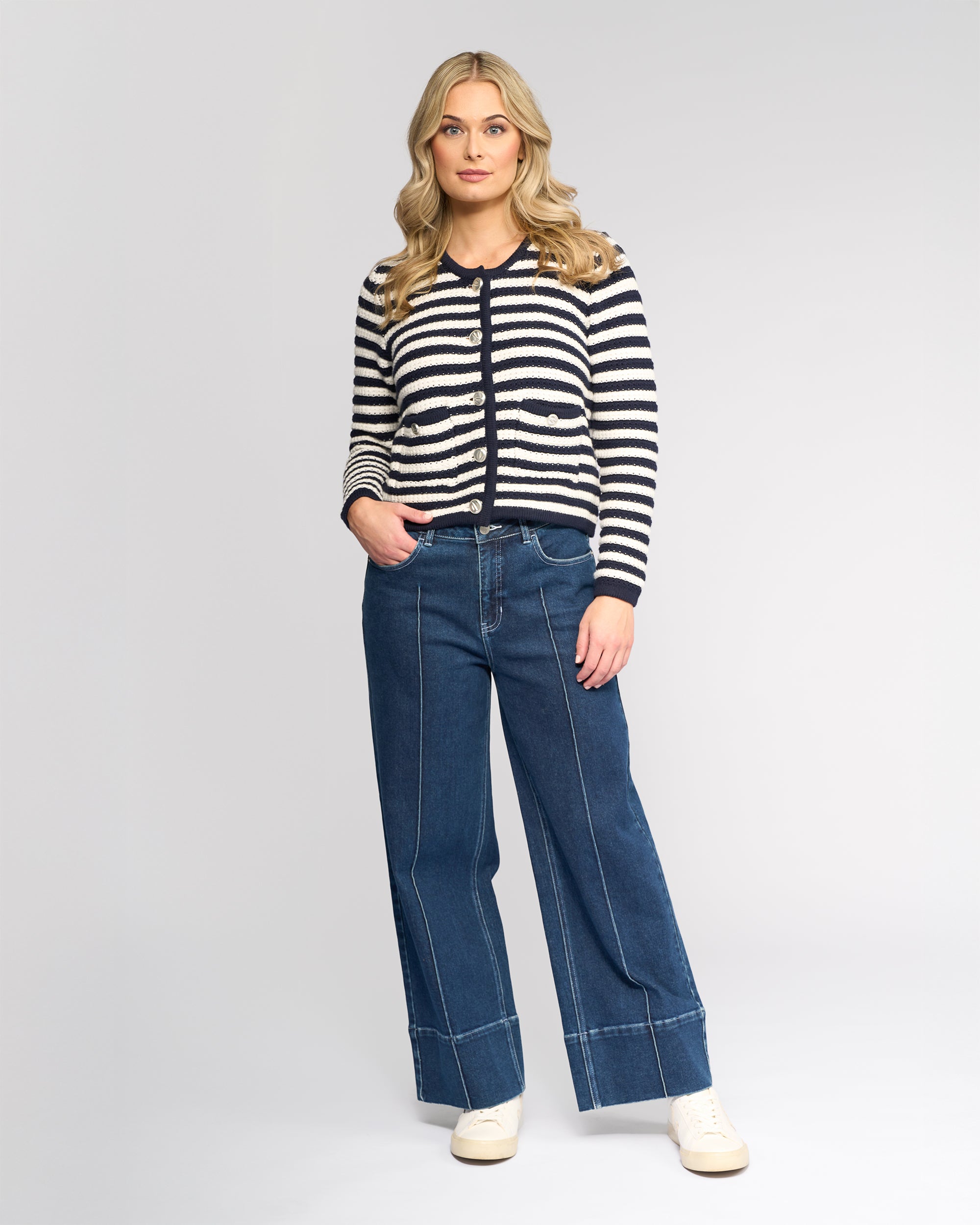 A woman with long blonde hair wears the Vassalli Avenue Cardi in Ink/Cream, styled with wide-leg blue jeans and white sneakers, standing against a plain light gray background.