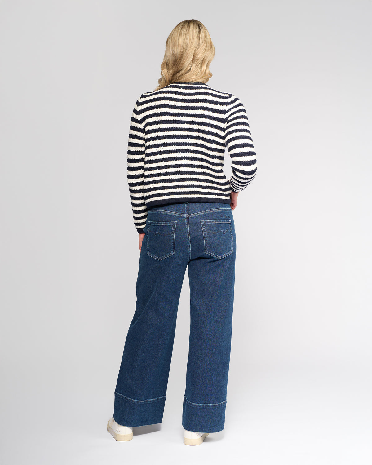 A person with shoulder-length blonde hair, wearing the Vassalli Avenue Cardi in Ink/Cream, wide-leg blue jeans, and white shoes stands facing away from the camera against a plain background.