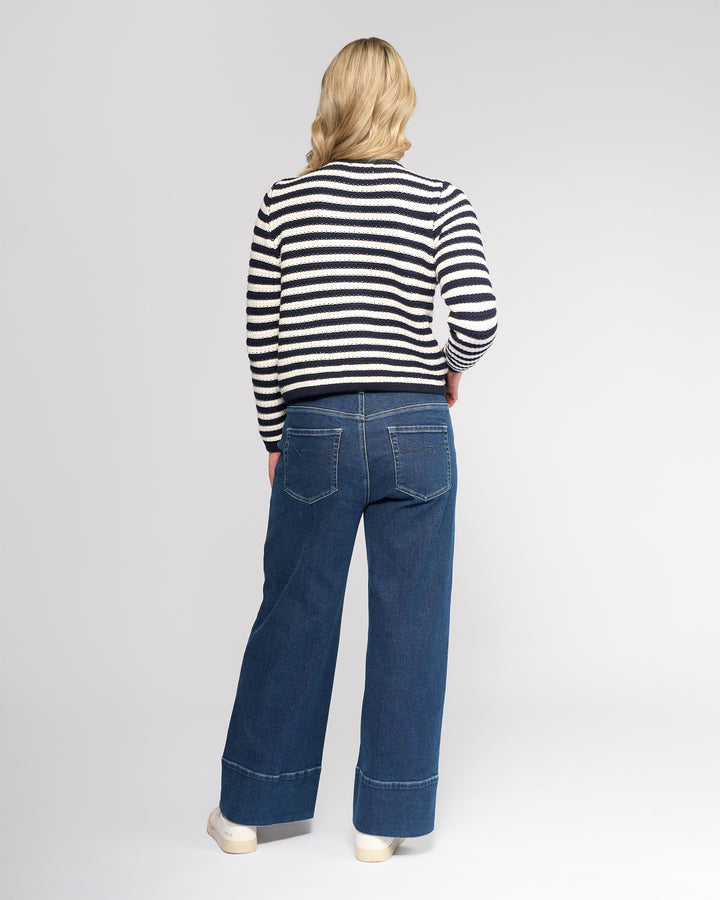 A person with shoulder-length blonde hair, wearing the Vassalli Avenue Cardi in Ink/Cream, wide-leg blue jeans, and white shoes stands facing away from the camera against a plain background.