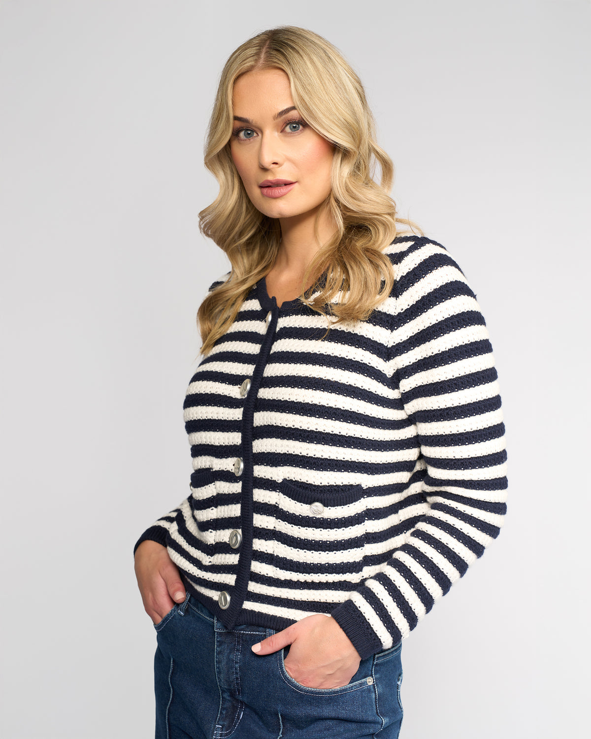 A woman with long blonde hair wears the Vassalli Avenue Cardi in Ink/Cream and blue jeans, standing with her hands in her pockets against a plain light background, looking at the camera.