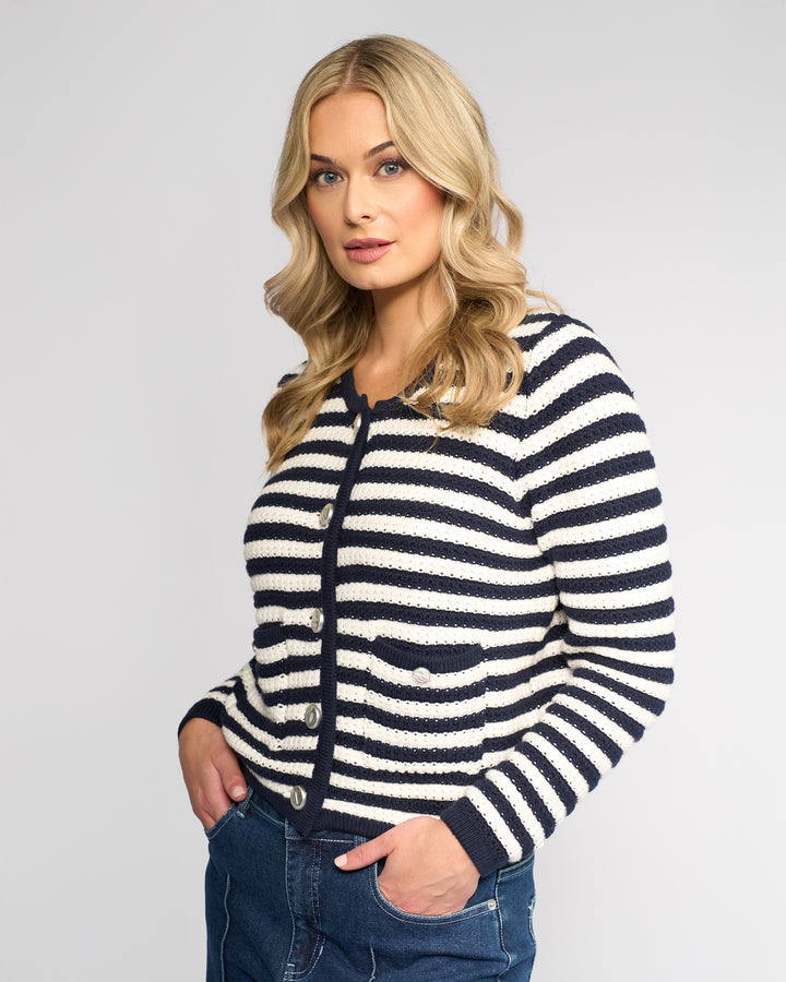 A woman with long blonde hair wears the Vassalli Avenue Cardi in Ink/Cream and blue jeans, standing with her hands in her pockets against a plain light background, looking at the camera.