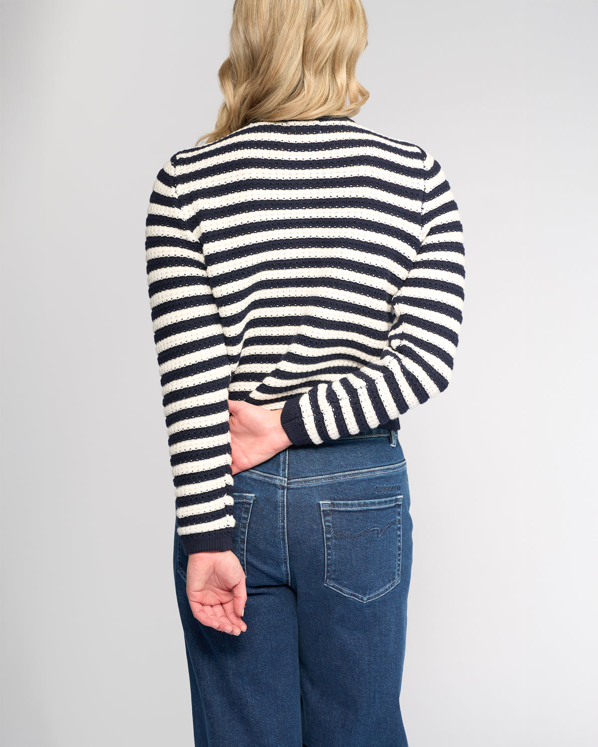 A person with blonde hair, seen from behind, wears the Vassalli Avenue Cardi - Ink/Cream, a black and white slim-fit cotton cardigan with smashed metallic buttons, paired with blue jeans. One arm is bent behind their back, holding the other at the elbow.