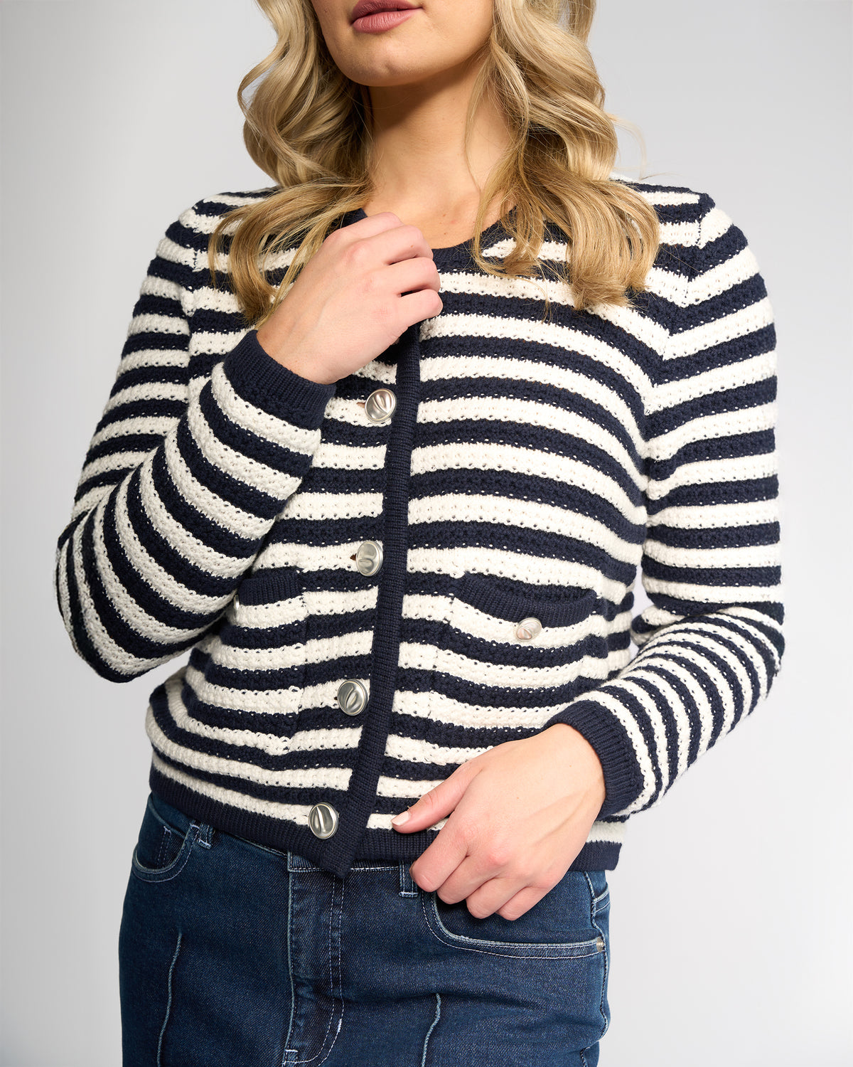 A woman with loose blonde waves stands against a plain light background, wearing the Vassalli Avenue Cardi - Ink/Cream with smashed metallic buttons and dark denim jeans, holding the edge of her black and white striped sweater.