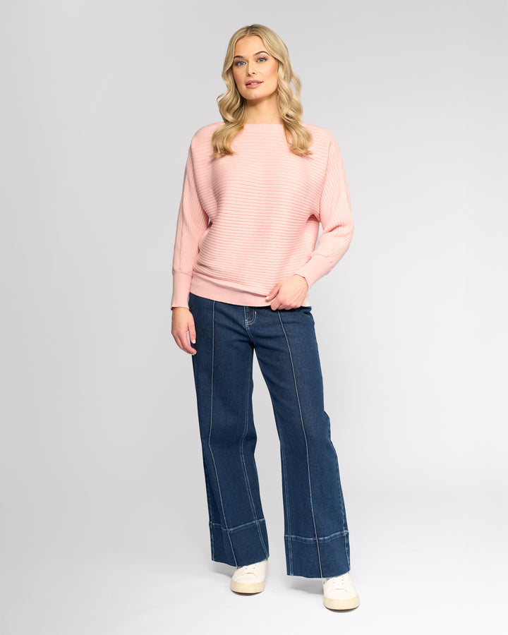 A woman with long blonde hair wears the Vassalli Kiki Sweater in Dusky Pink, styled with dark blue wide-leg jeans featuring white stitching and white sneakers, standing against a light gray background.