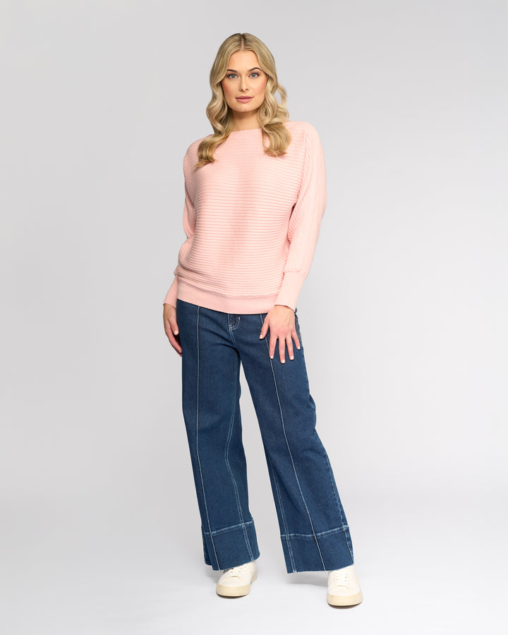 A woman with long blonde hair stands against a light gray background, wearing the Vassalli Kiki Sweater in dusky pink, paired with wide-leg blue jeans featuring white stitching and white sneakers. She faces forward with a relaxed expression.