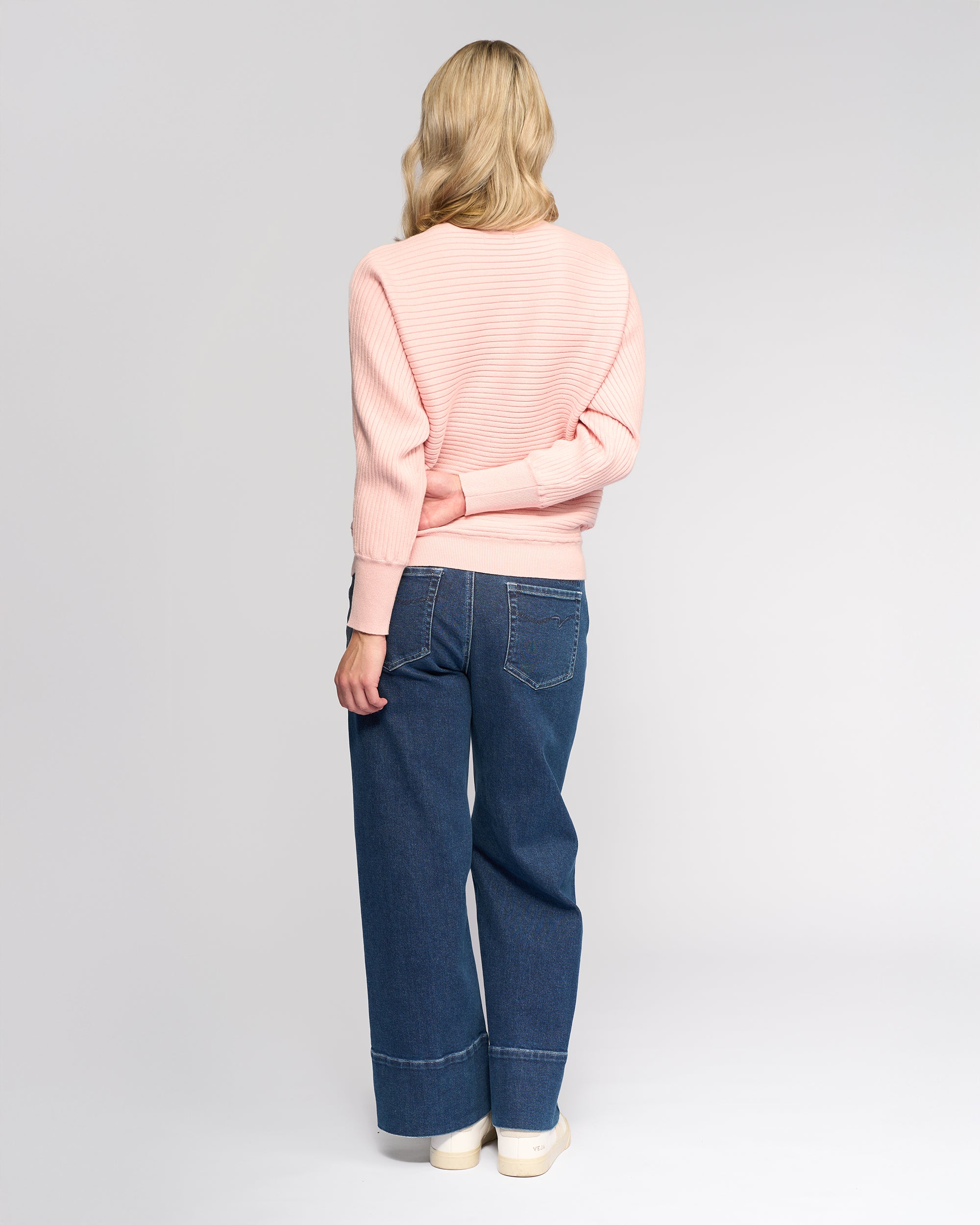 A person with long blonde hair wears the Vassalli Kiki Sweater in dusky pink, wide-leg blue jeans, and white shoes, standing facing away from the camera with one arm behind their back.