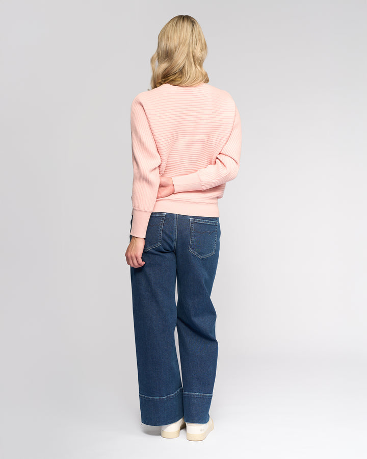 A person with long blonde hair wears the Vassalli Kiki Sweater in dusky pink, wide-leg blue jeans, and white shoes, standing facing away from the camera with one arm behind their back.