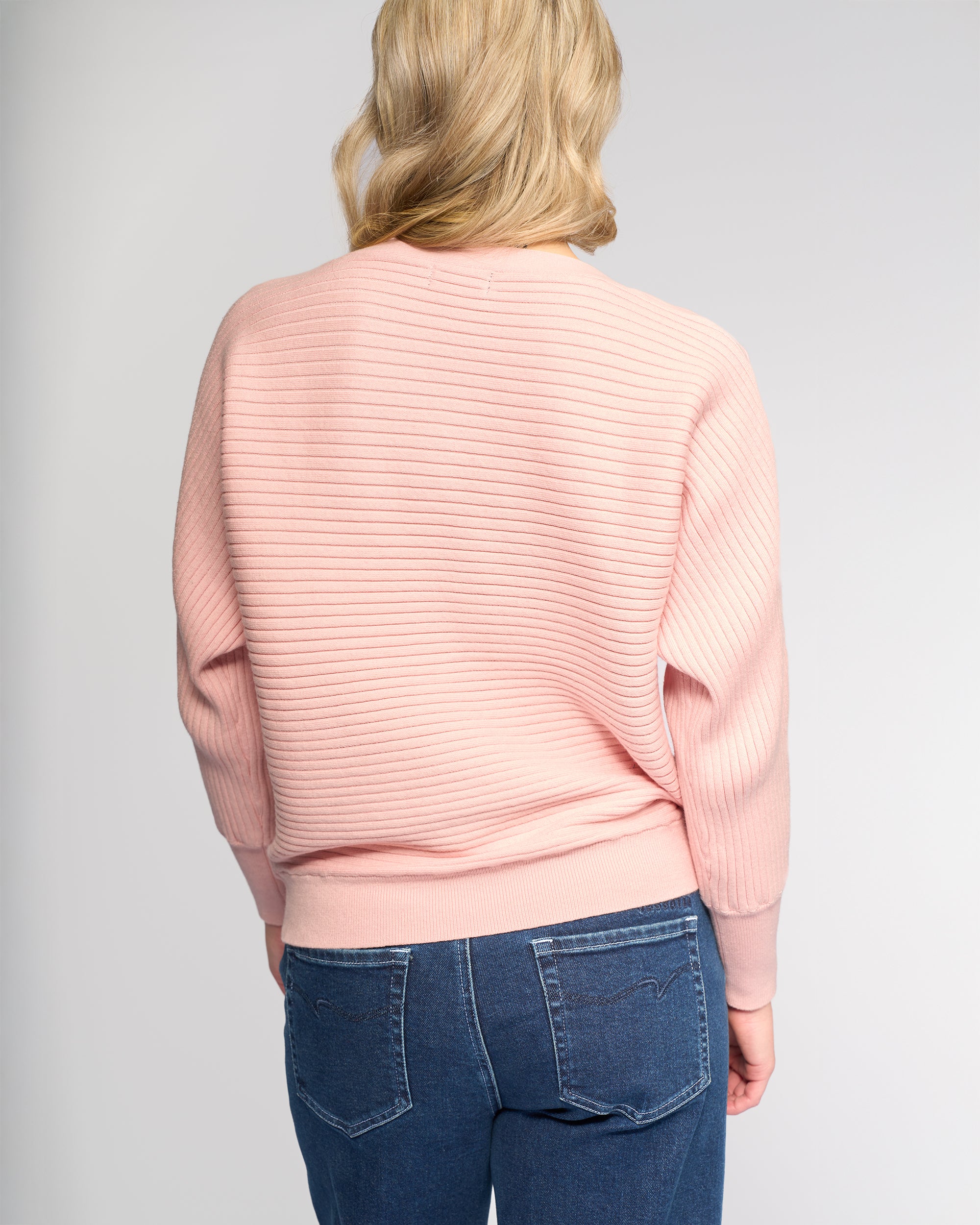 A person with blonde hair is seen from behind, wearing the Vassalli Kiki Sweater in dusky pink and blue jeans against a plain light background.