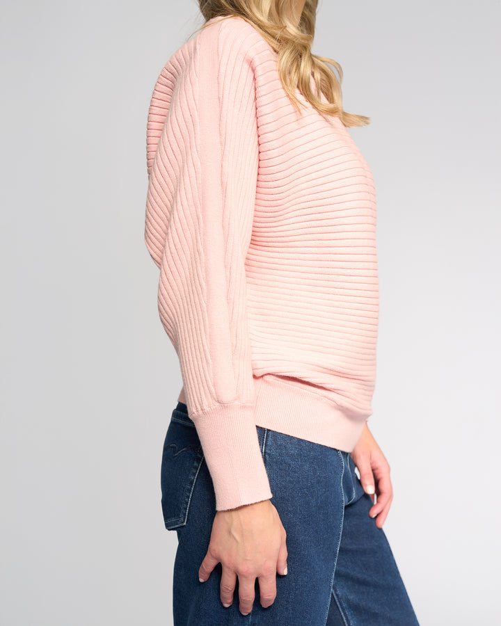 A person is shown from head to upper legs wearing the Vassalli Kiki Sweater in dusky pink with long sleeves and blue jeans, standing sideways against a plain light background.