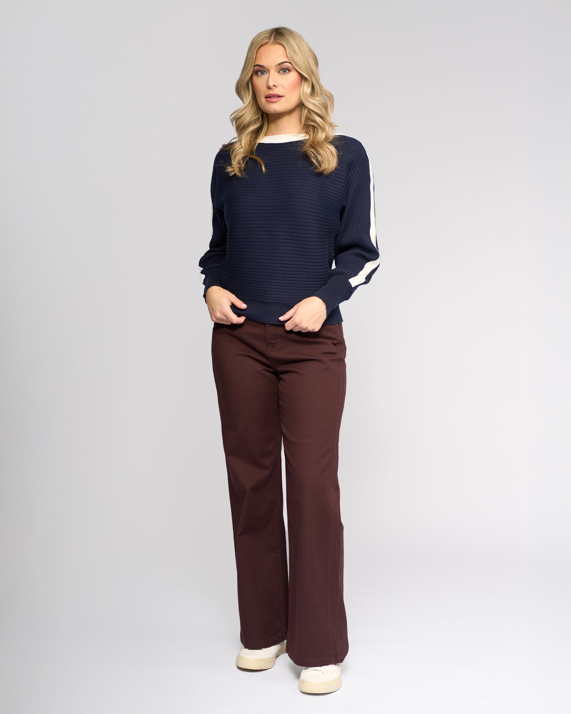 A woman with long blonde hair stands facing forward, wearing the Vassalli Kiki Sweater in Ink/Winter White, featuring batwing sleeves with white stripes, brown wide-leg pants, and cream shoes against a plain light gray background.