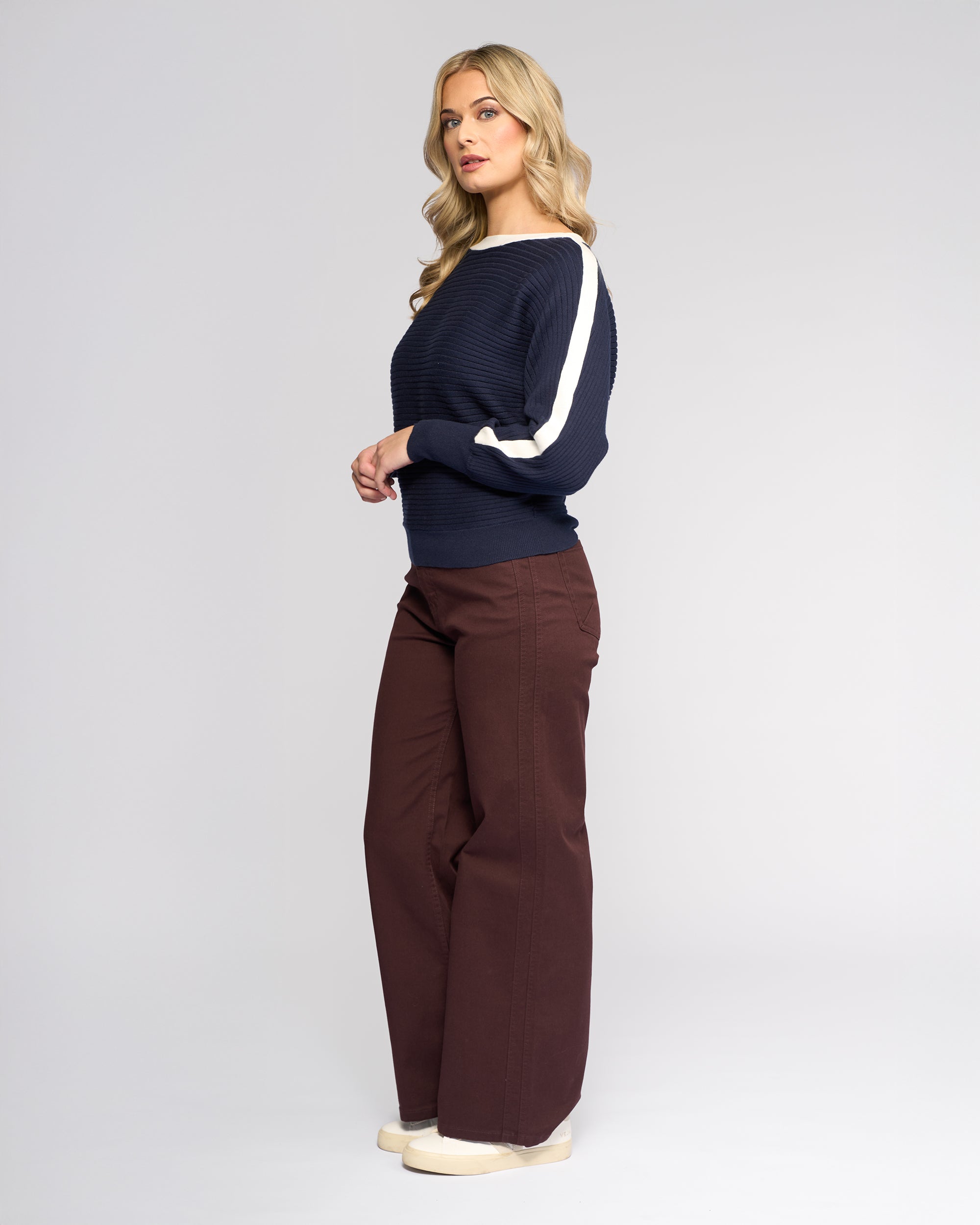 A woman with long blonde hair stands sideways against a light gray background, wearing the Vassalli Kiki Sweater in Ink/Winter White with white sleeve stripes, brown wide-leg pants, and beige shoes.