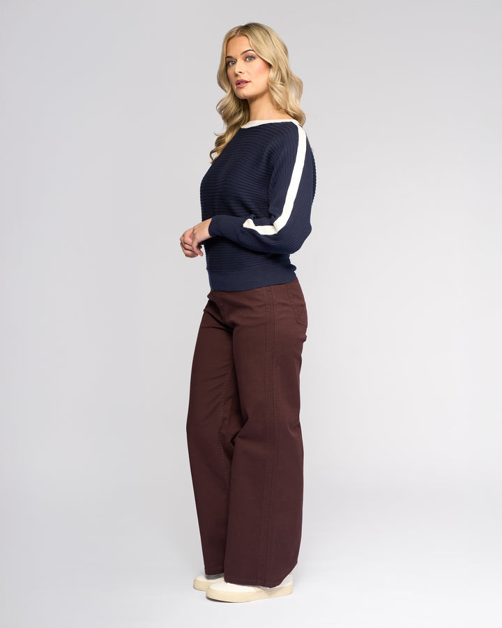 A woman with long blonde hair stands sideways against a light gray background, wearing the Vassalli Kiki Sweater in Ink/Winter White with white sleeve stripes, brown wide-leg pants, and beige shoes.