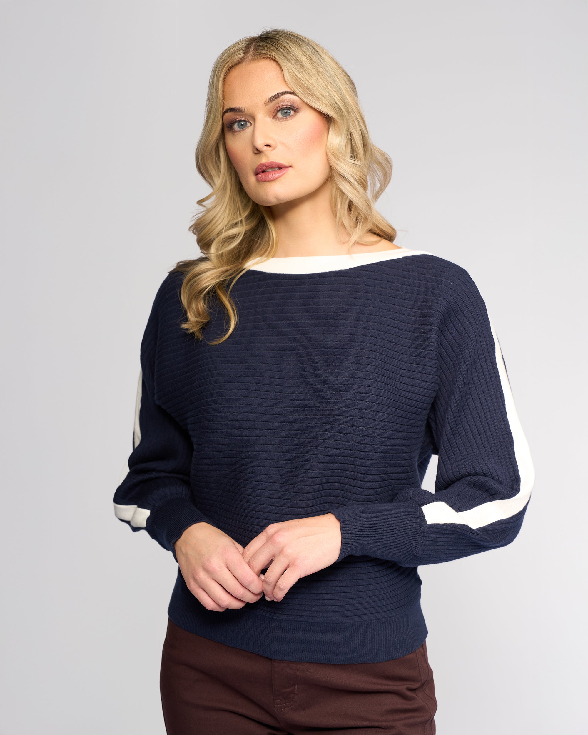 A woman with long blonde hair wears the Vassalli Kiki Sweater in Ink/Winter White, a relaxed-fit ribbed knit with white sleeve stripes, paired with brown pants, standing against a plain light gray background.