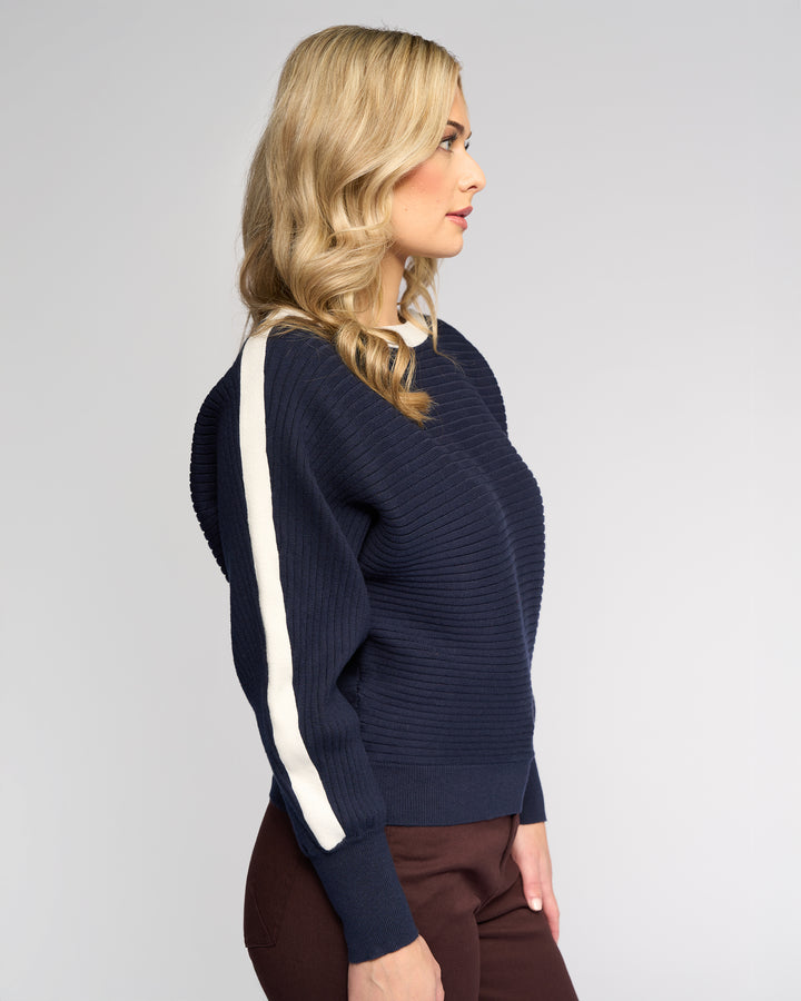 A woman with wavy blonde hair models the Vassalli Kiki Sweater in Ink/Winter White, a relaxed-fit knit with batwing sleeves and white sleeve stripes, paired with brown pants against a plain light gray backdrop.