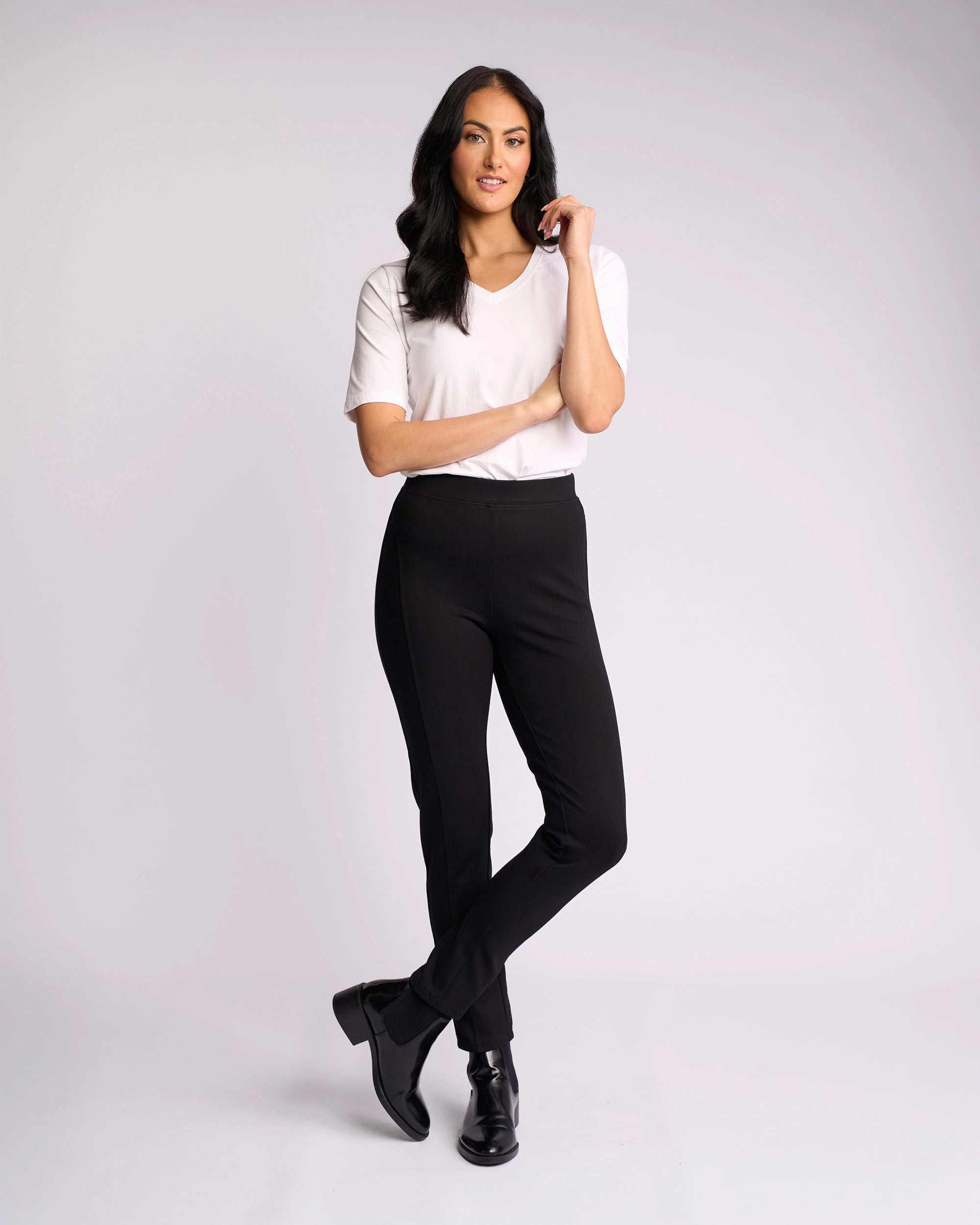 A woman with long dark hair stands against a plain background, wearing a white short-sleeve top and Vassalli Fortune Pant - Black. She crosses her legs, touches her hair, and smiles gently.