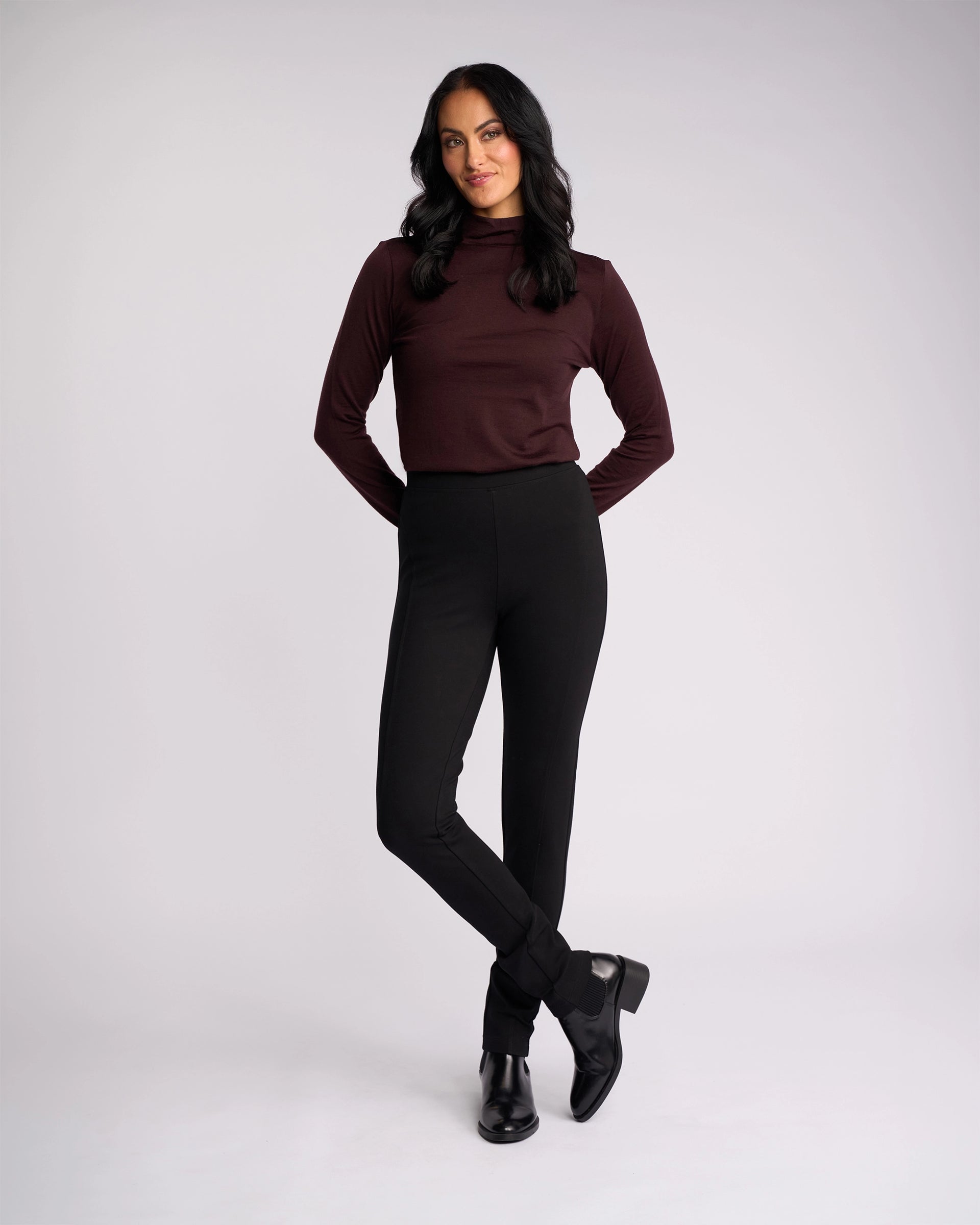 A woman with long dark hair stands against a plain background, wearing the Vassalli Toll Ponti Pant in black and a maroon turtleneck. She smiles slightly, hands behind her back, looking forward.
