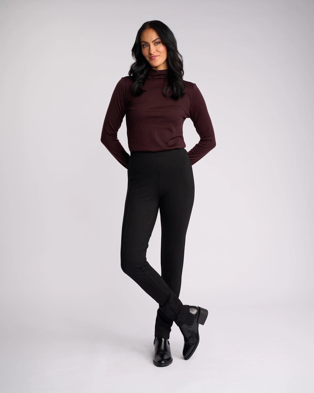 A woman with long dark hair stands against a plain background, wearing the Vassalli Toll Ponti Pant in black and a maroon turtleneck. She smiles slightly, hands behind her back, looking forward.