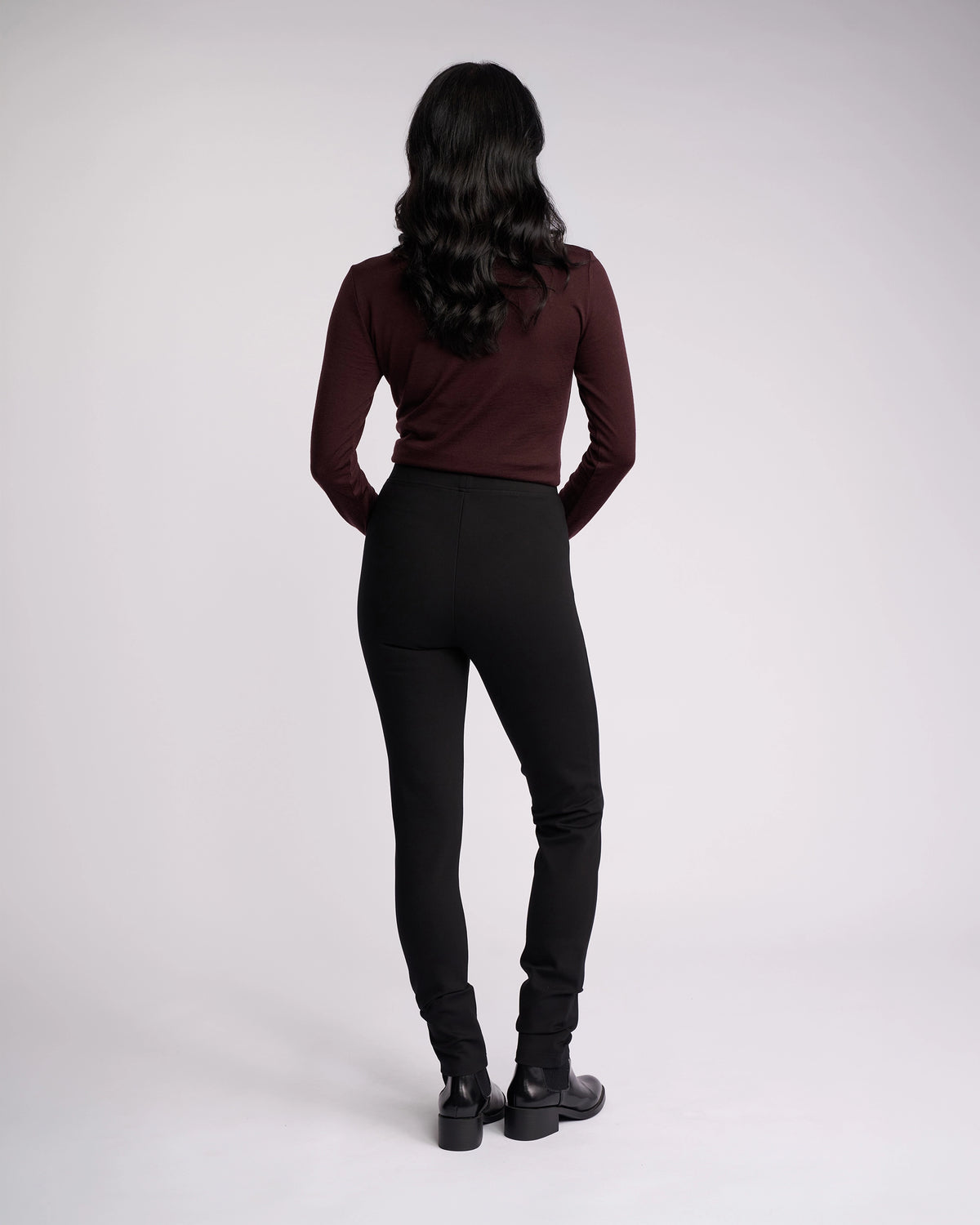 A person with long, wavy dark hair faces away from the camera, wearing a maroon long-sleeve top and Vassalli's Toll Ponti Pant in black, featuring a high-rise skinny leg, paired with black boots against a plain light background.