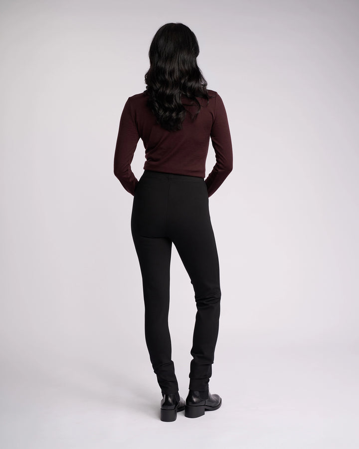 A person with long, wavy dark hair faces away from the camera, wearing a maroon long-sleeve top and Vassalli's Toll Ponti Pant in black, featuring a high-rise skinny leg, paired with black boots against a plain light background.