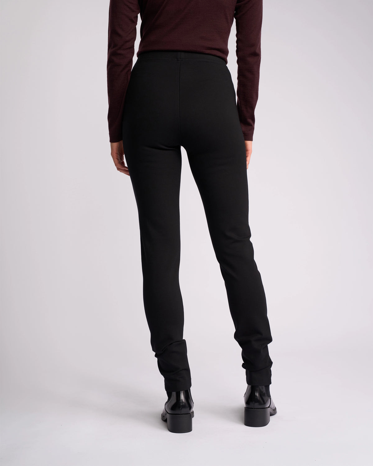 A person stands facing away from the camera, wearing a long-sleeve dark top, Vassalli Toll Ponti Pant - Black, and black ankle boots against a plain light background.