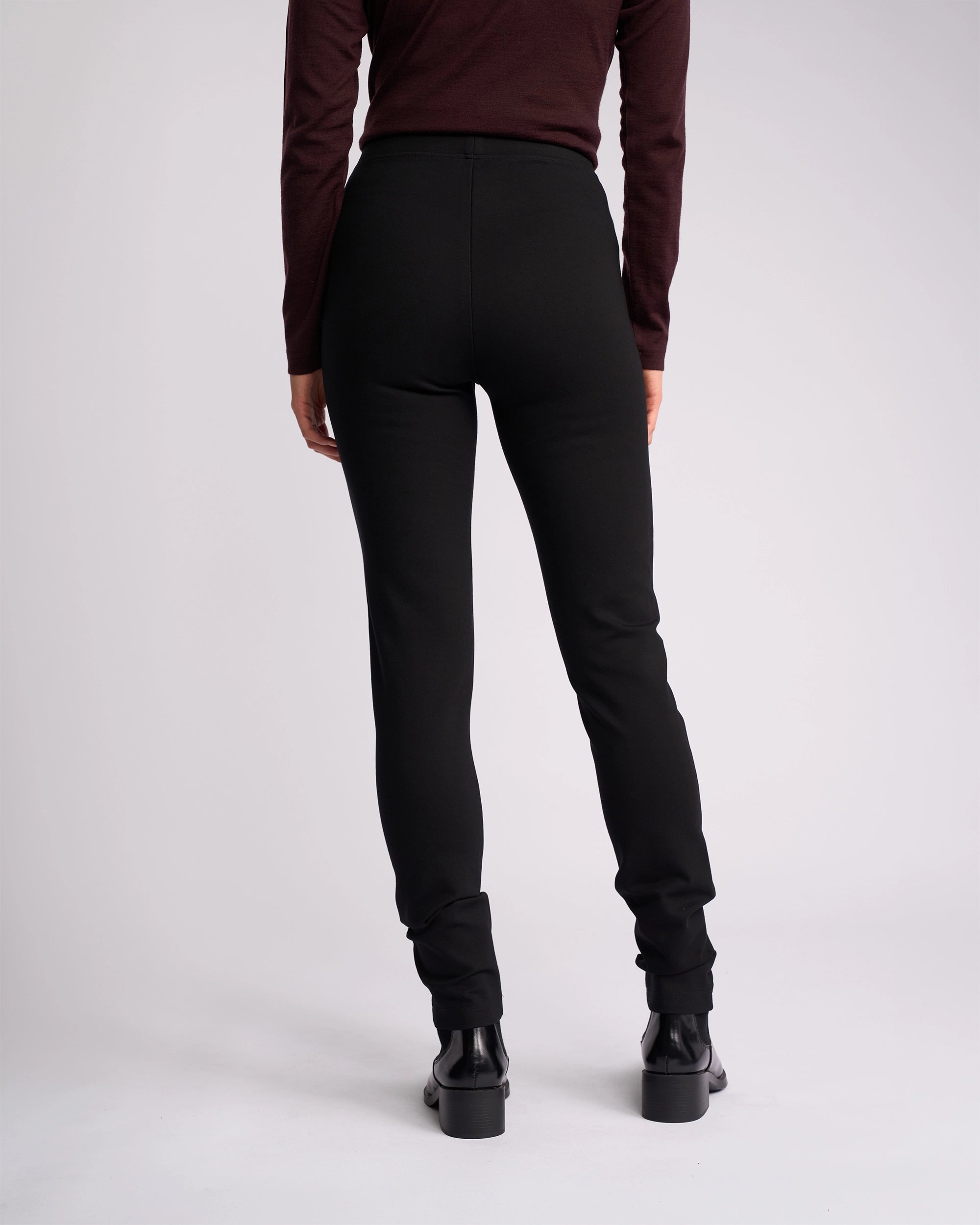 A person stands facing away from the camera, wearing a long-sleeve dark top, Vassalli Toll Ponti Pant - Black, and black ankle boots against a plain light background.