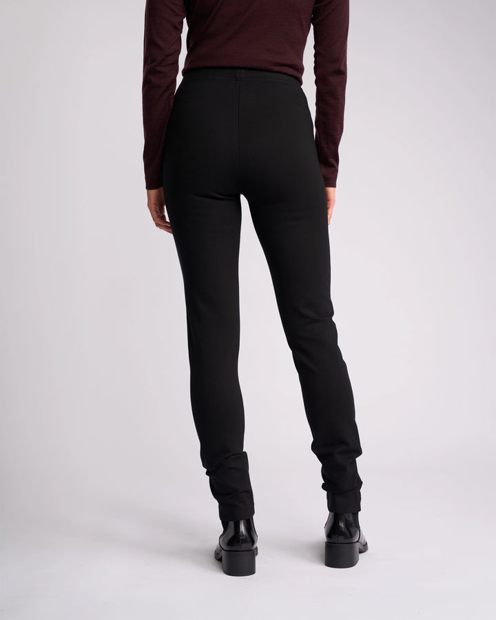 A person stands facing away from the camera, wearing a long-sleeve dark top, Vassalli Toll Ponti Pant - Black, and black ankle boots against a plain light background.