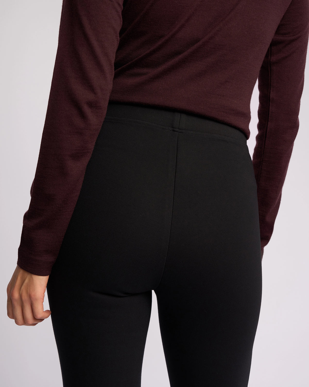 A person is shown from the back wearing Vassalli’s Toll Ponti Pant in black, paired with a long-sleeved maroon top, their left arm relaxed at their side against a plain light background.