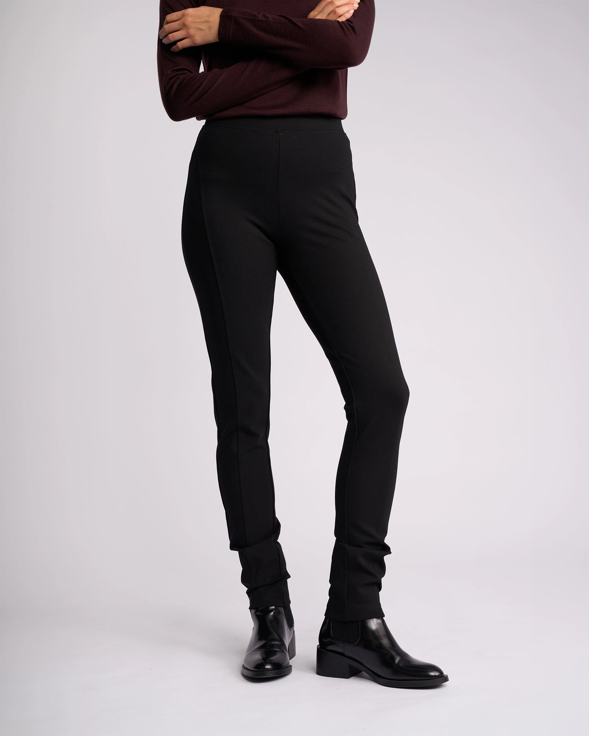 A person with arms crossed models the Vassalli Toll Ponti Pant in black, paired with a long-sleeve dark top and black ankle boots, against a plain light background. The individual's head is not shown in the image.