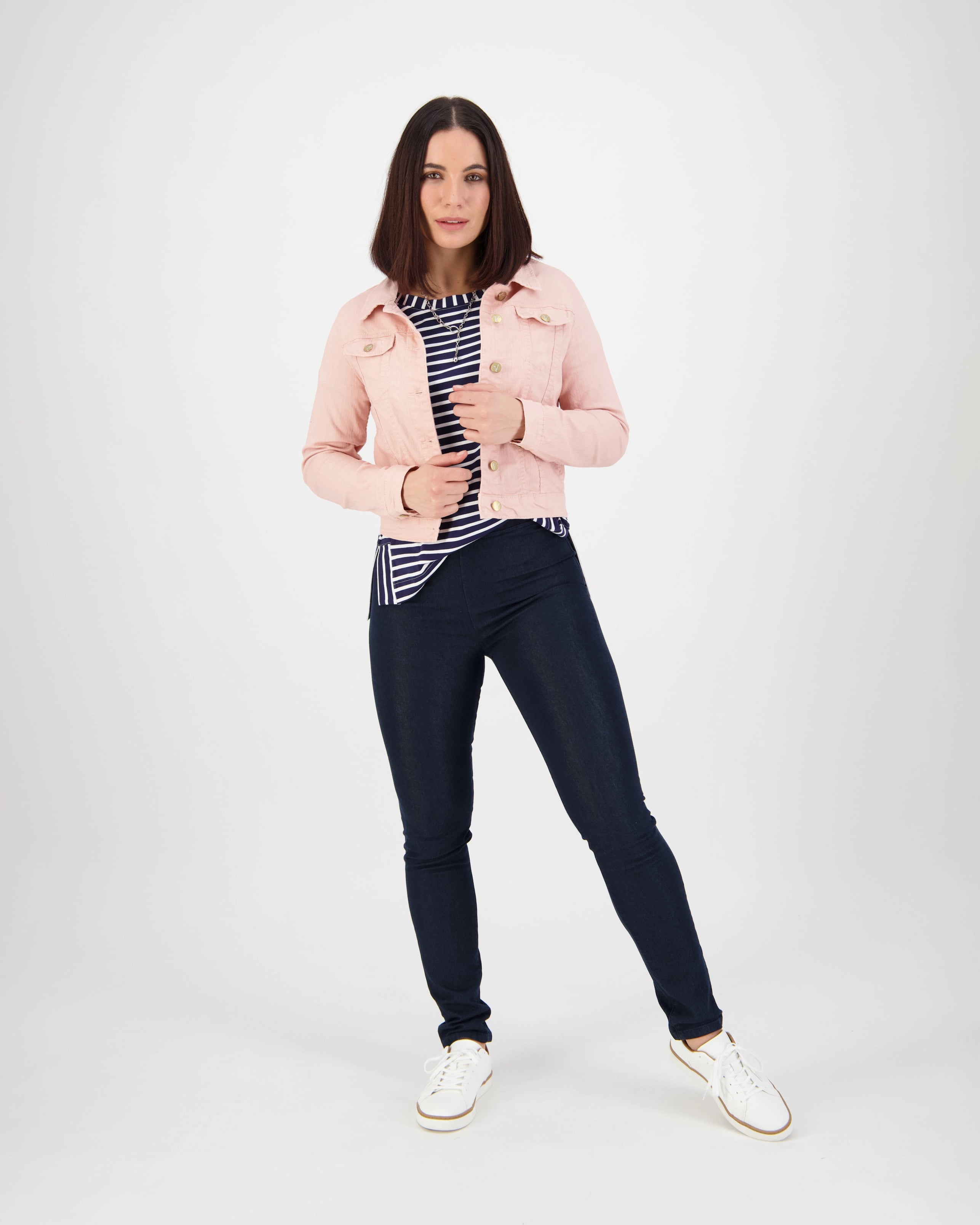 A woman stands against a white background wearing the Vassalli Sloane Denim Jacket in French Rose over a black and white striped top, dark skinny jeans, and white sneakers. She looks forward holding her jacket.