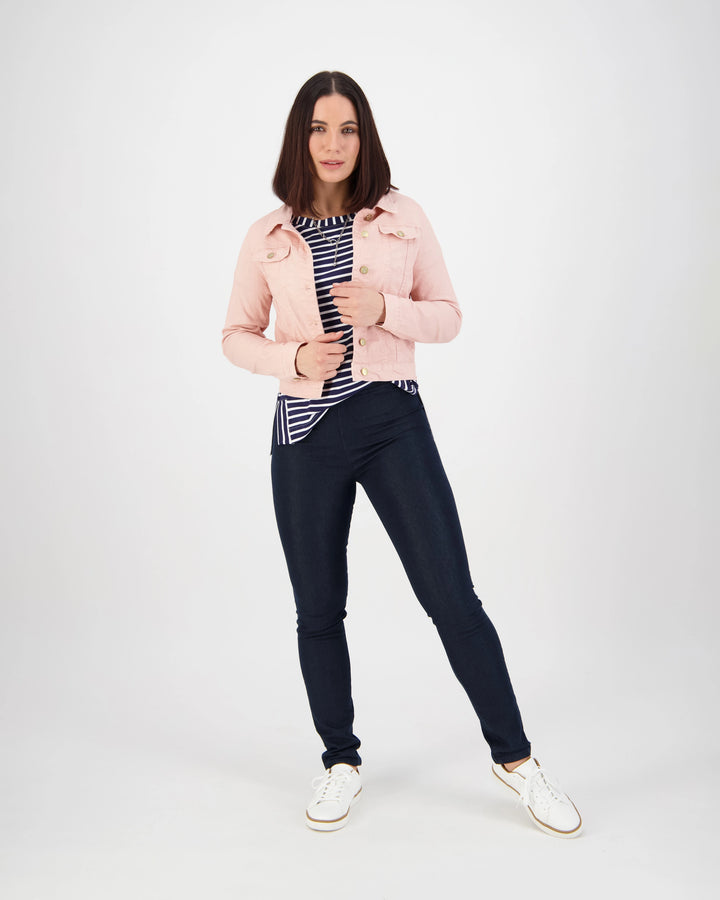 A woman stands against a white background wearing the Vassalli Sloane Denim Jacket in French Rose over a black and white striped top, dark skinny jeans, and white sneakers. She looks forward holding her jacket.