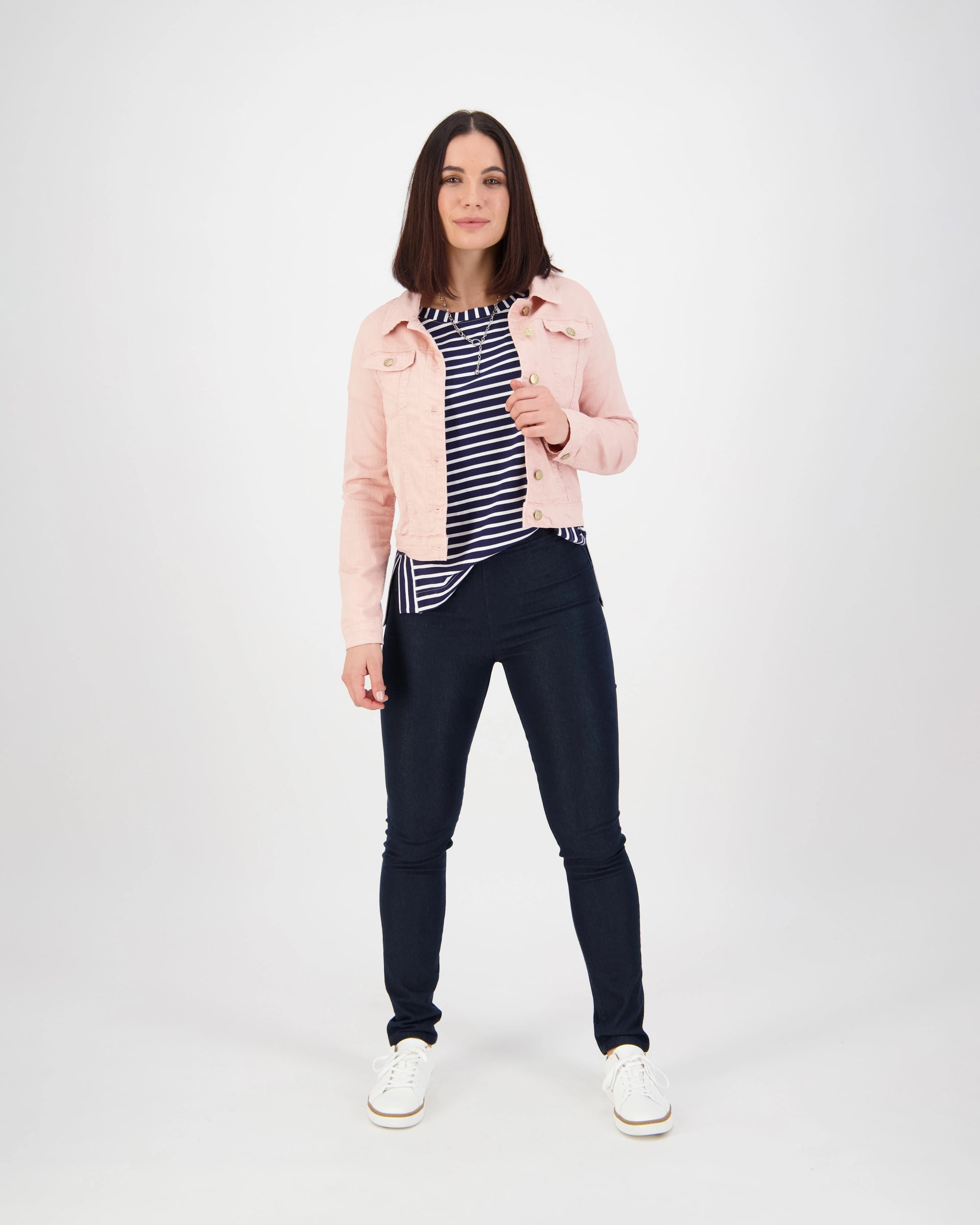 A woman with shoulder-length brown hair wears the Vassalli Sloane Denim Jacket in French Rose over a navy and white striped shirt, paired with dark skinny jeans and white sneakers. She faces forward with a neutral expression.