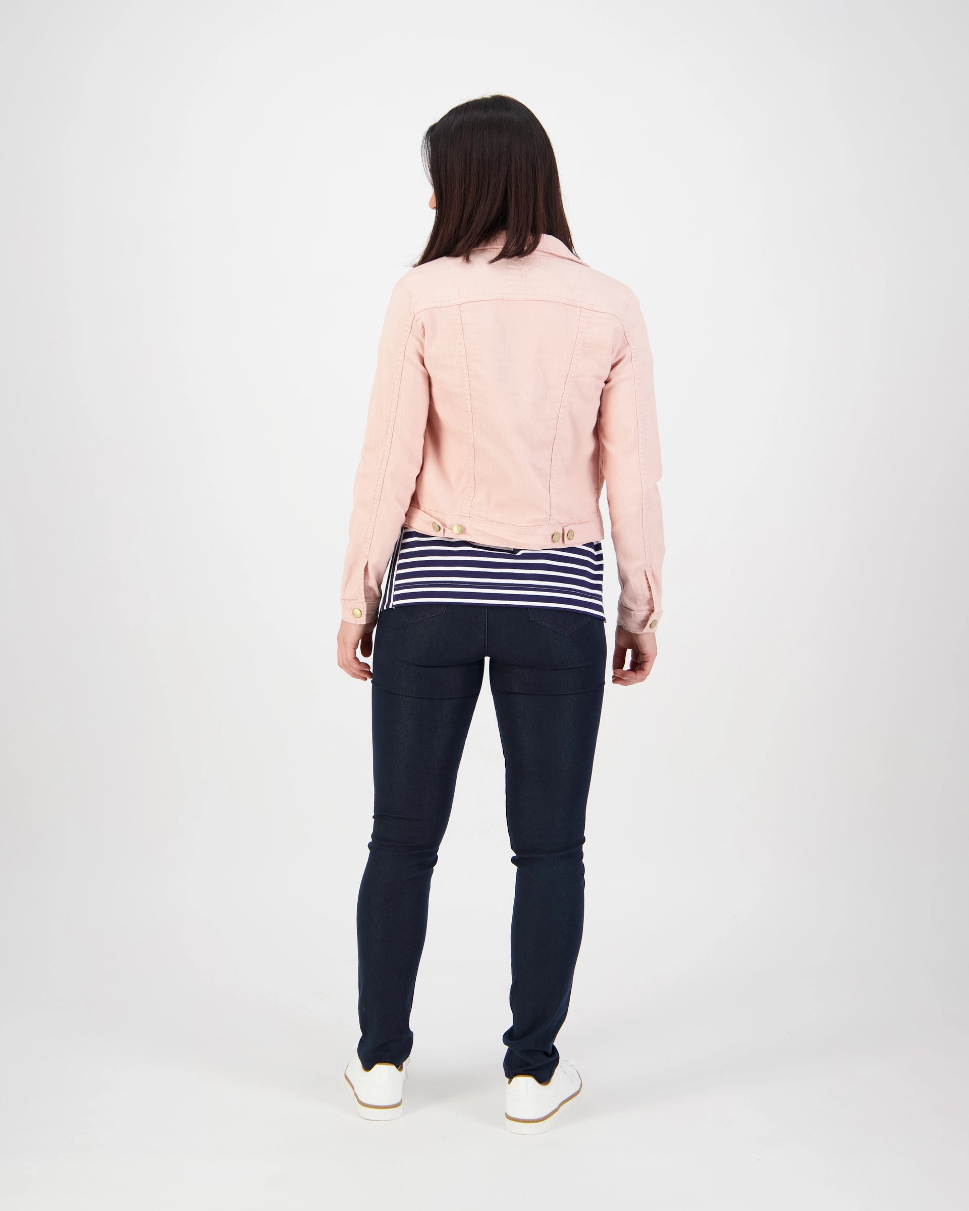 A woman stands facing away from the camera, wearing the Vassalli Sloane Denim Jacket in French Rose, a navy and white striped shirt, dark skinny jeans, and white shoes against a plain white background.