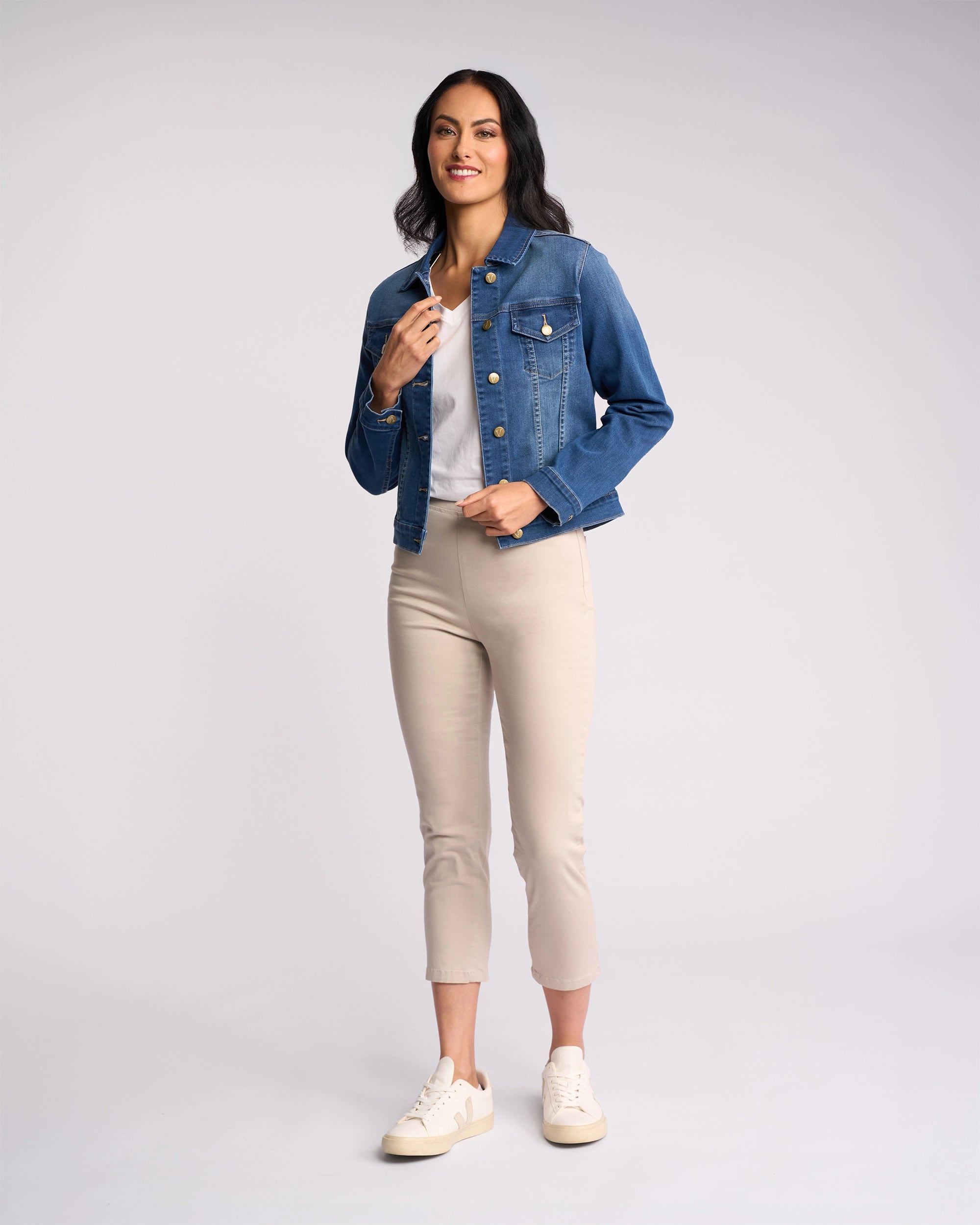 A woman smiles in a studio, wearing the Vassalli Ratio Jacket in blue wash denim, paired with a white shirt, beige cropped pants, and white sneakers against a plain light background.