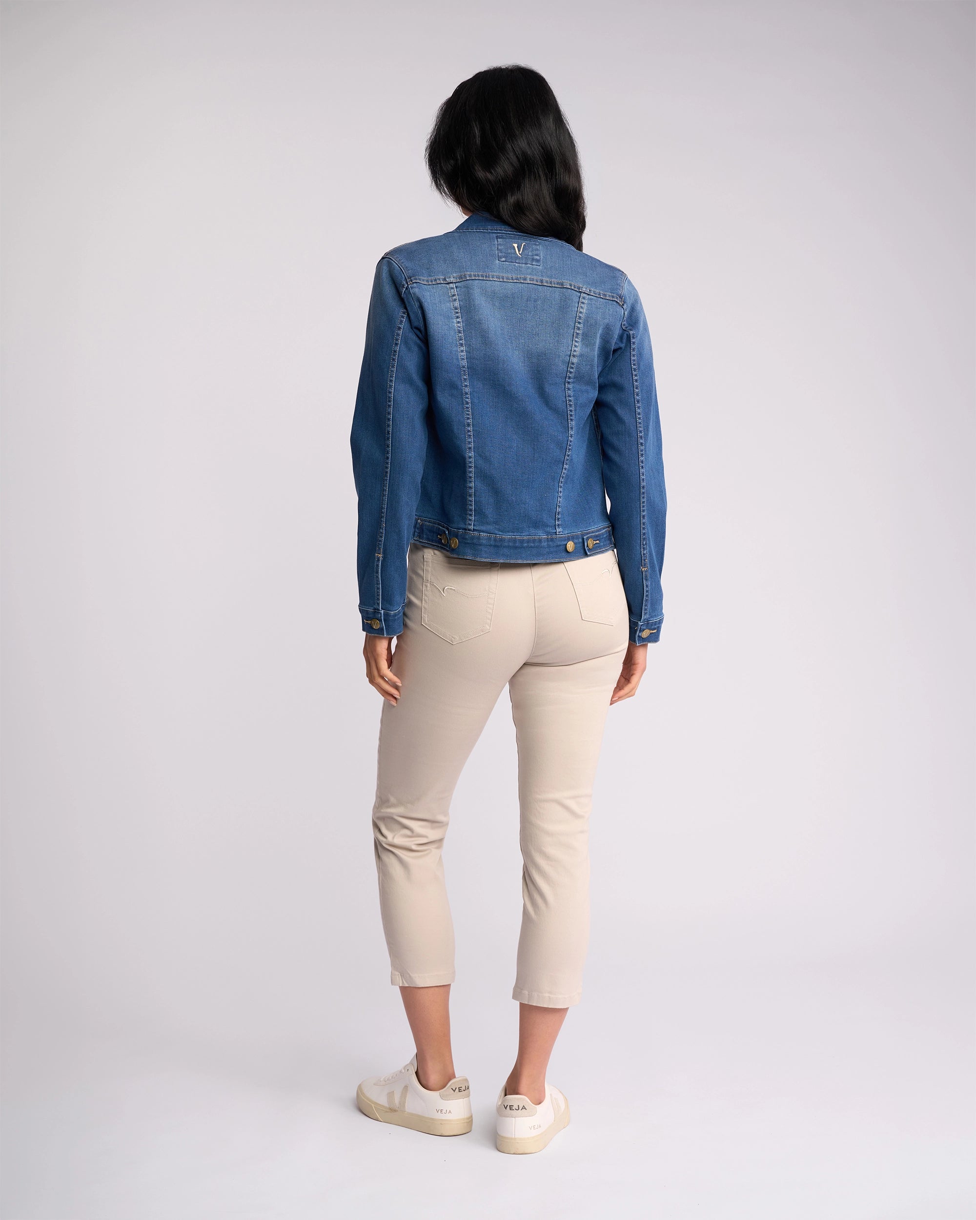 A woman faces away from the camera wearing the Vassalli Ratio Jacket in Blue Wash Denim, paired with light beige cropped pants and white sneakers, set against a plain light background.