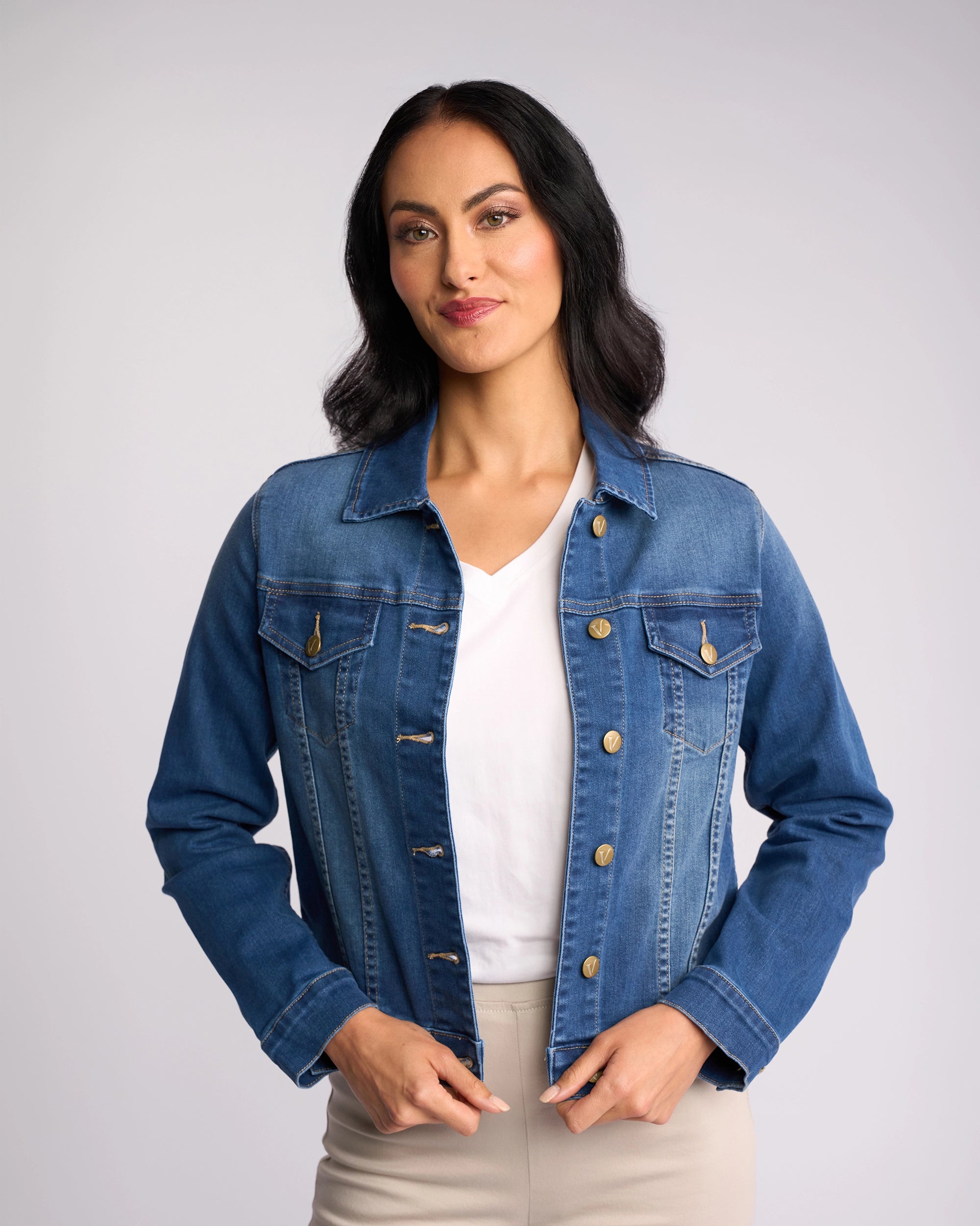 A woman with dark hair wears the Vassalli Ratio Jacket in blue wash denim, styled relaxed over a white shirt and beige pants, standing against a light background and smiling softly as she holds the jacket.