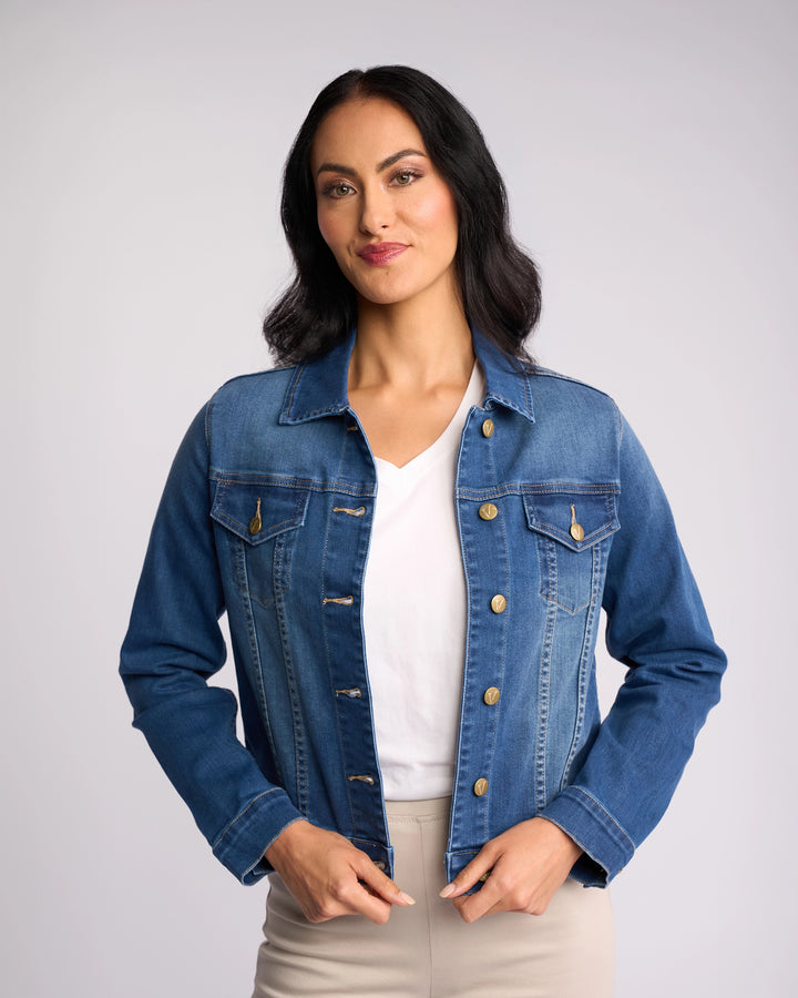A woman with dark hair wears the Vassalli Ratio Jacket in blue wash denim, styled relaxed over a white shirt and beige pants, standing against a light background and smiling softly as she holds the jacket.