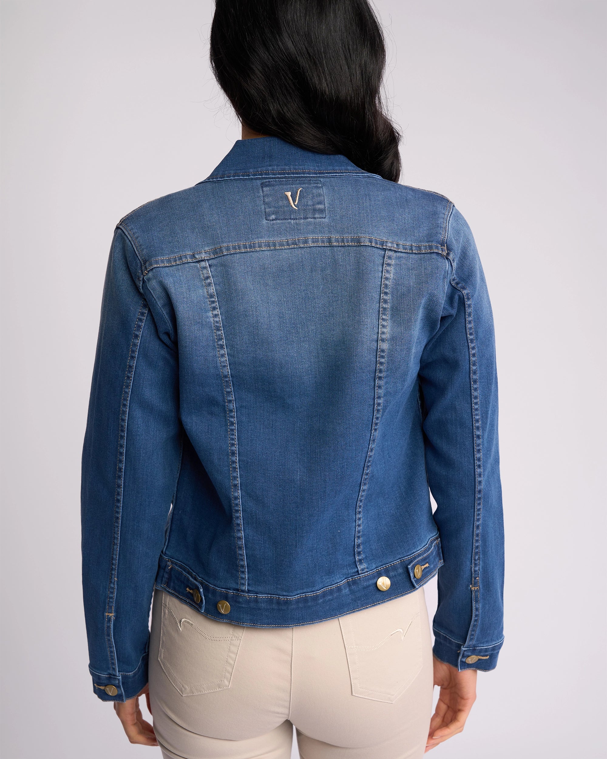 A woman with long dark hair is seen from the back wearing the Vassalli Ratio Jacket in Blue Wash Denim and relaxed-fit light beige pants, standing against a plain light background.
