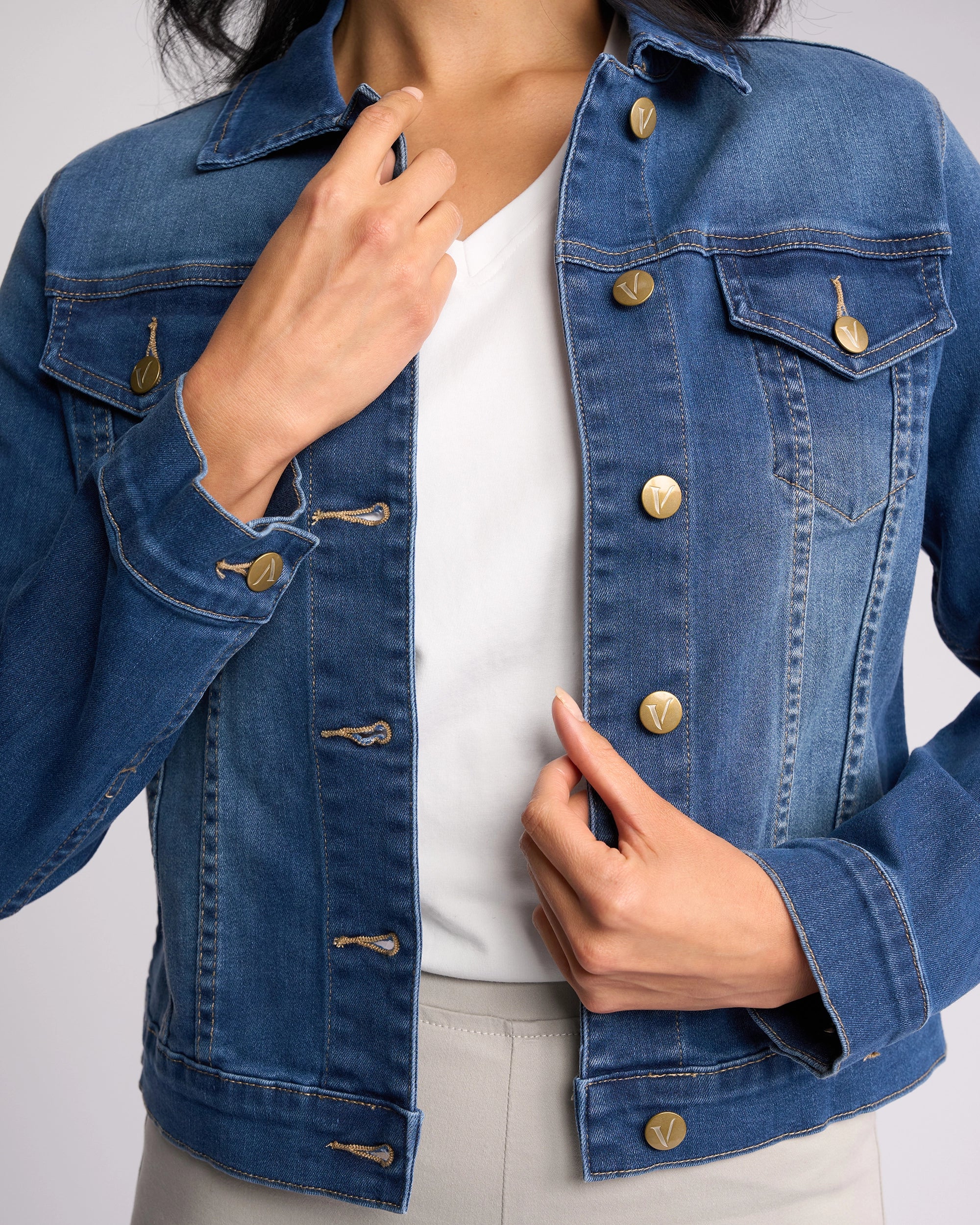 A person is buttoning the Vassalli Ratio Jacket - Blue Wash Denim over a white shirt, with one hand by the collar and the other by the bottom buttons. Their face is not visible.